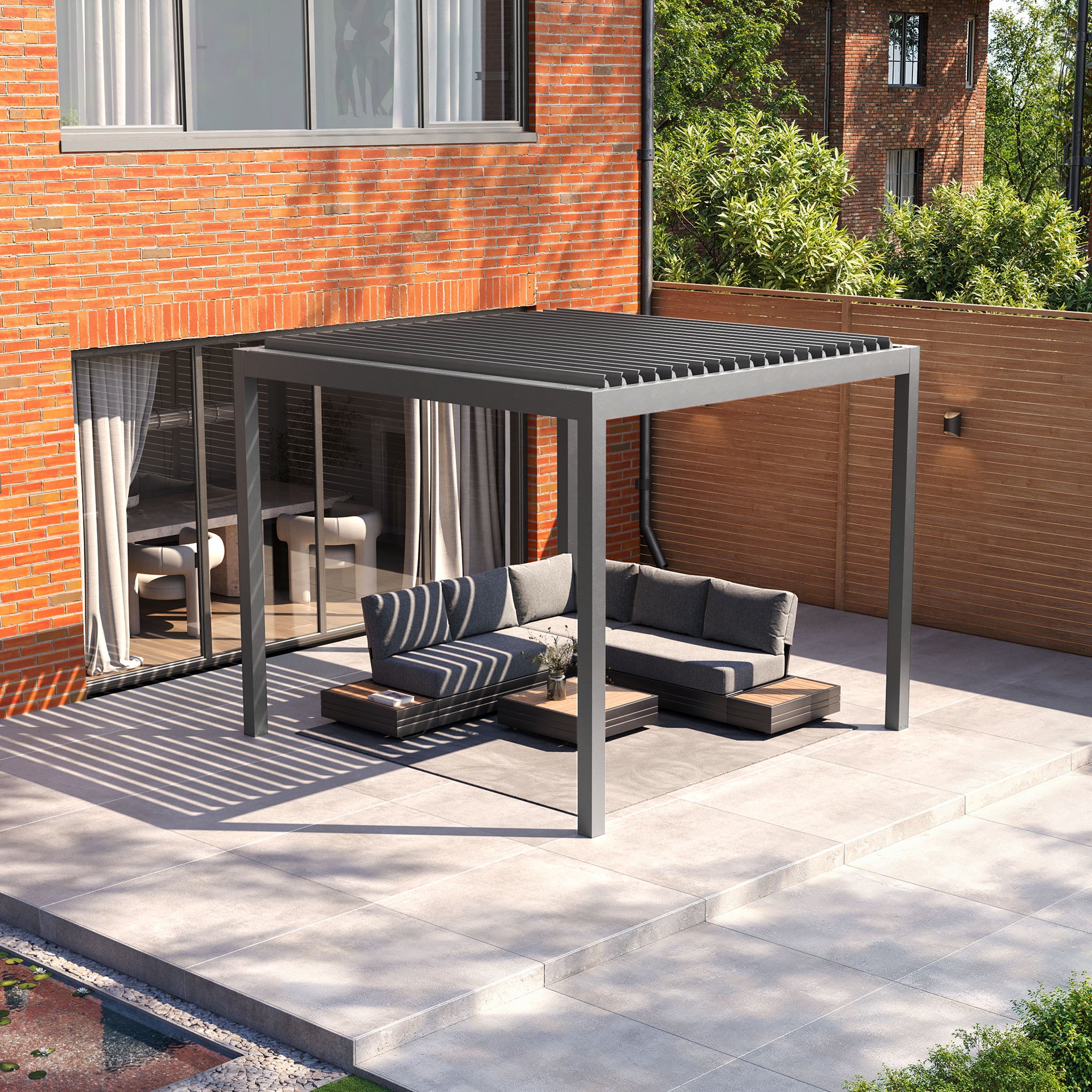 A modern patio features a PergoSTET Deluxe 3m x 3m grey pergola with motorized LED louvres over a sectional sofa and wooden table on a tiled terrace. The LED lighting enriches the ambiance at dusk, making evenings inviting outside the brick building.