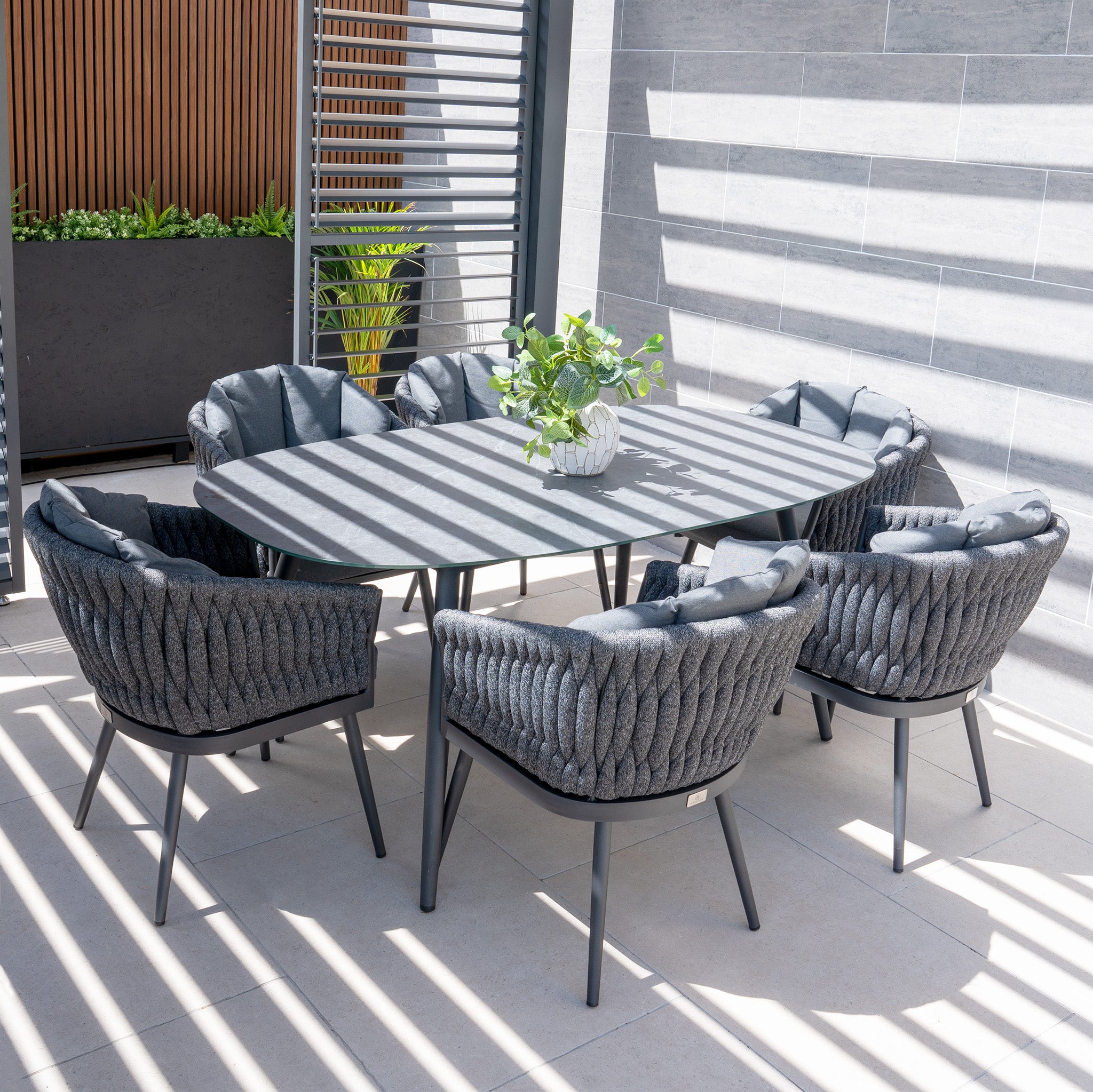 The Palma 6 Seat Rope Oval Dining Set with Ceramic Table in Grey includes six comfortable, UV-resistant chairs surrounding an elegant oval table on a tiled patio, shaded by slatted panels.