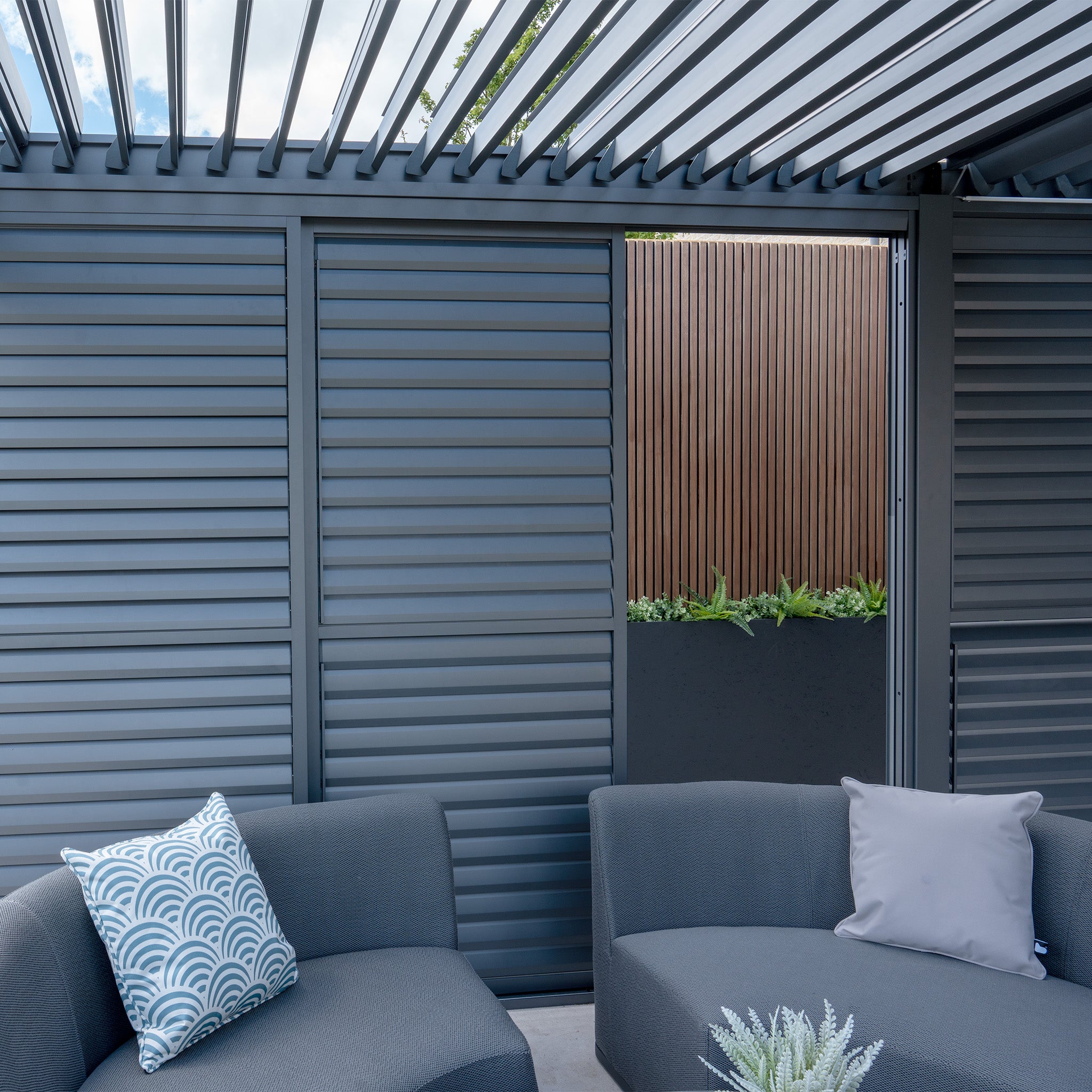 Contemporary patio featuring gray seating, striped accent pillows, and PergoSTET Deluxe Louvred Sliding Doors 3m Side in Grey, with a vertical wooden fence backdrop.