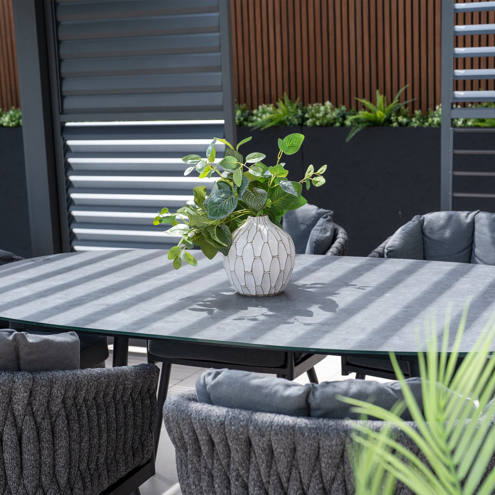 The Palma 6 Seat Rope Oval Dining Set with Ceramic Table in Grey is elegantly arranged with a white vase and green plant, encircled by gray cushioned chairs showcasing Harbour Lifestyle Palma's UV-resistant design and durable aluminum frames, all set against modern paneling.