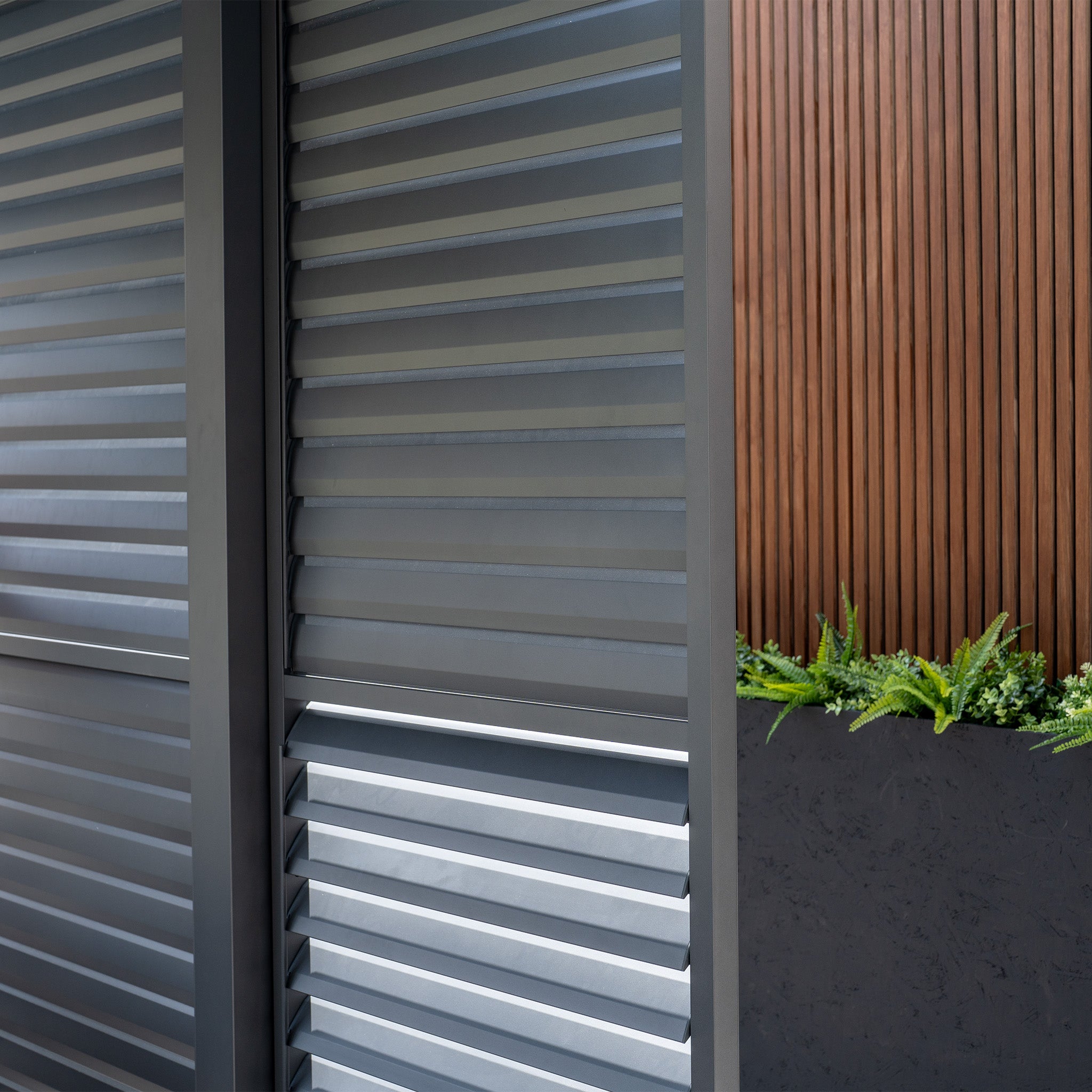 PergoSTET Deluxe Louvred Sliding Doors 3m Side in Grey beside a planter with green plants against vertical wooden slats.