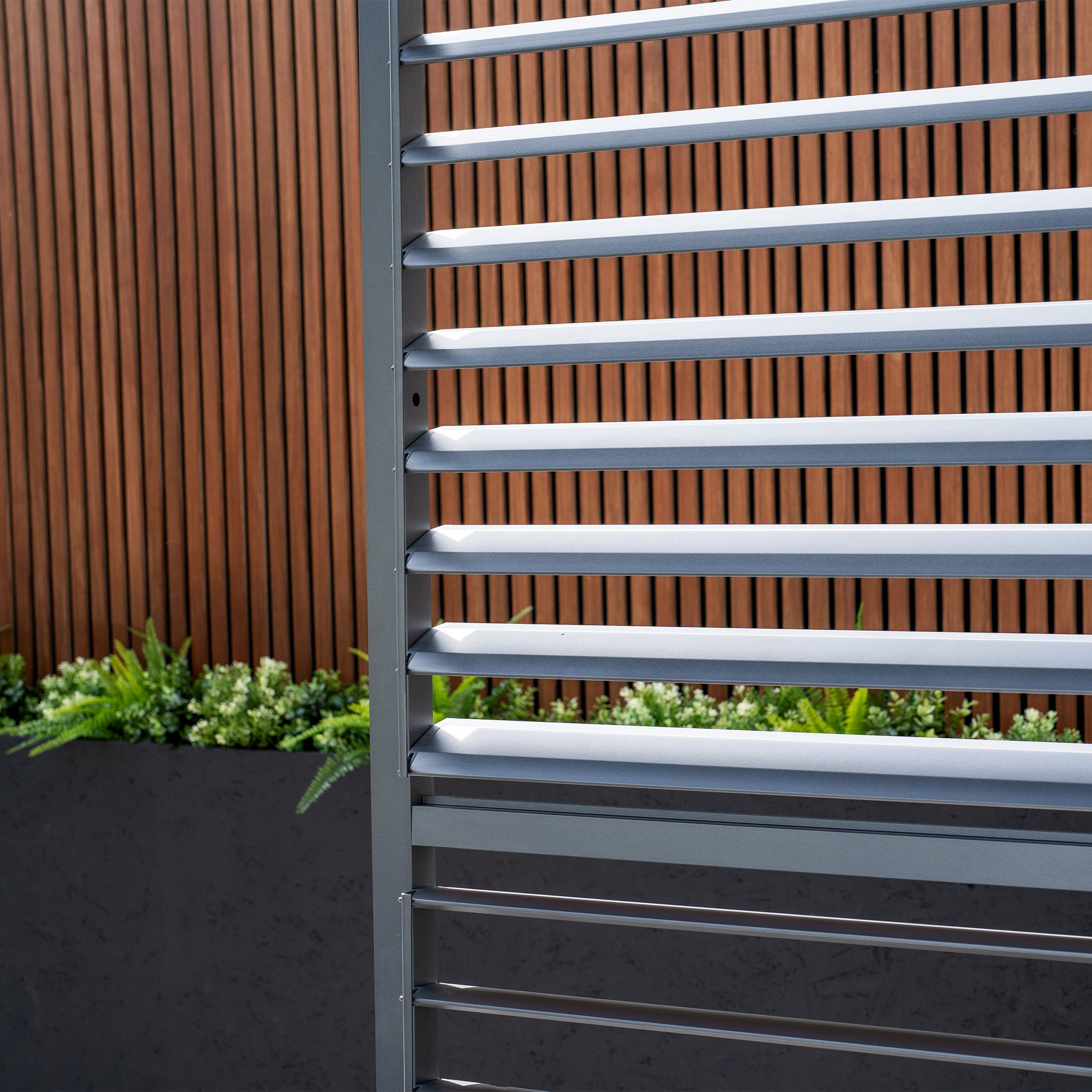 A close-up showcases the PergoSTET Deluxe Louvred Side Panel for 3m Side in Grey, set against a wooden wall and lush green plants, creating an elegant contrast.