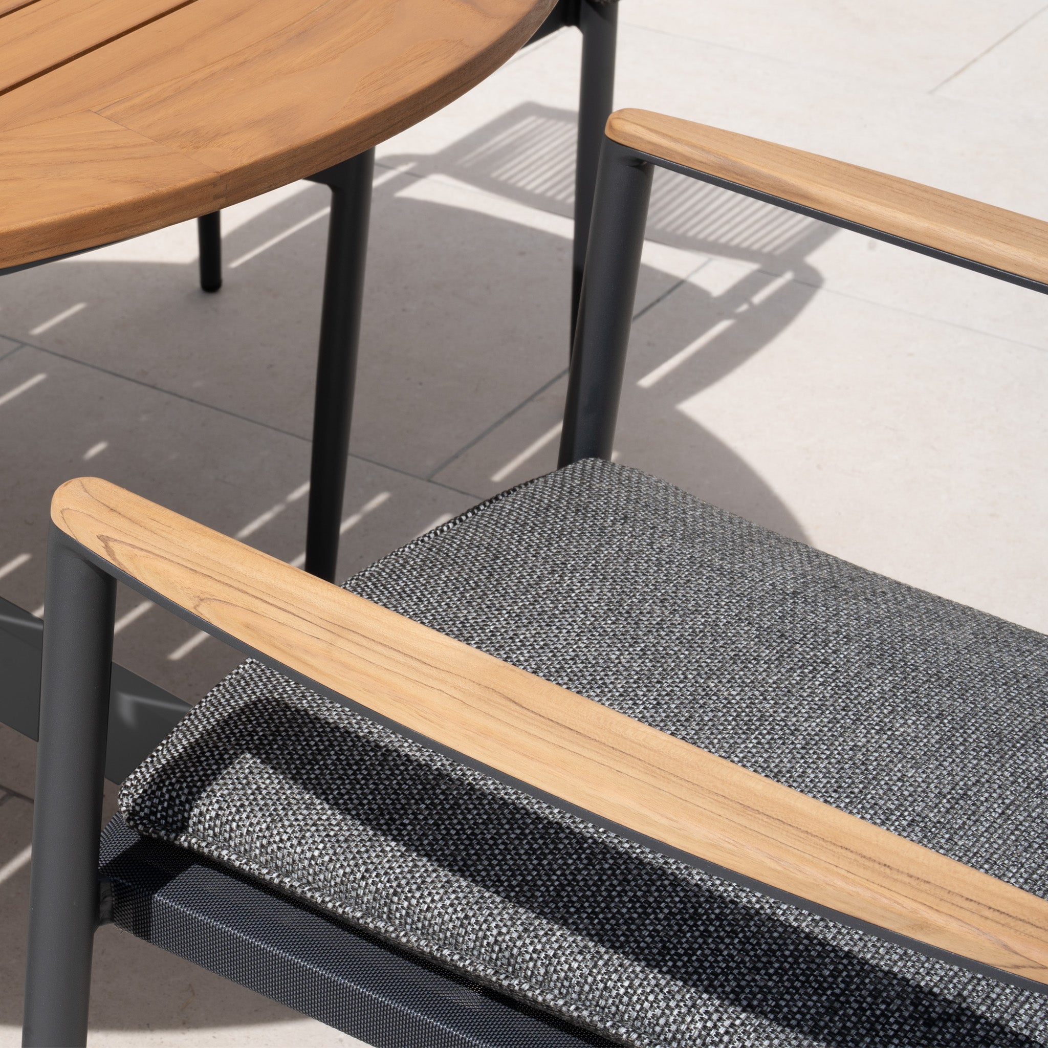 Close-up of the Tellaro 4 Seat Round Dining Set with a teak table in charcoal, featuring a wooden chair with a gray cushioned seat, on a sunlit patio for an inviting dining experience.