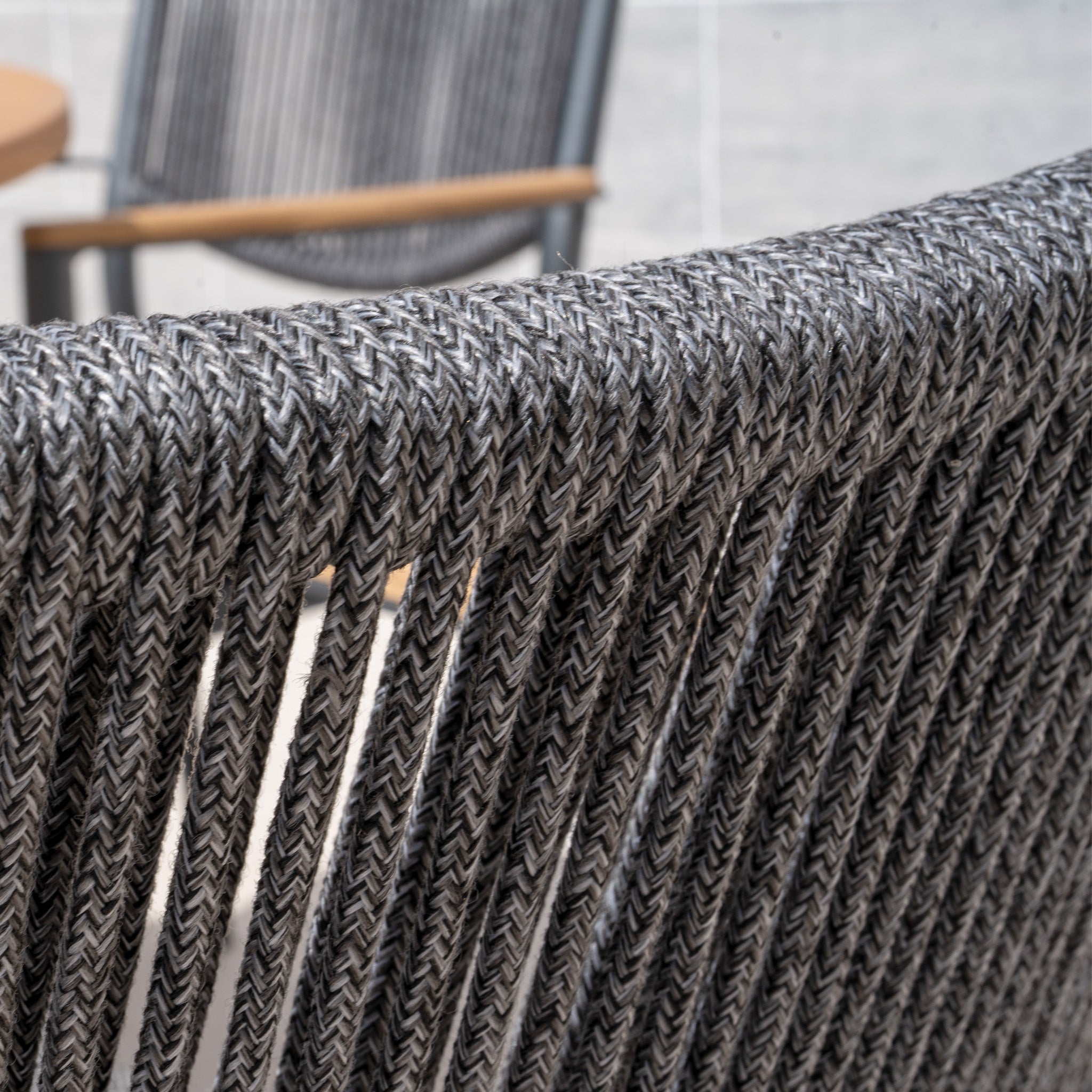 Close-up of a woven gray rope chair from the Tellaro 4 Seat Round Dining Set with Teak Table in Charcoal, with another chair blurred in the background.