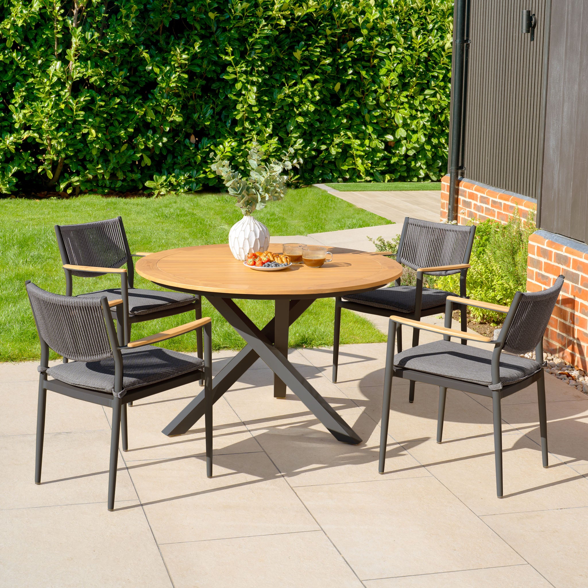 The Tellaro 4 Seat Round Dining Set with a teak table in charcoal is showcased on a patio surrounded by lush greenery.