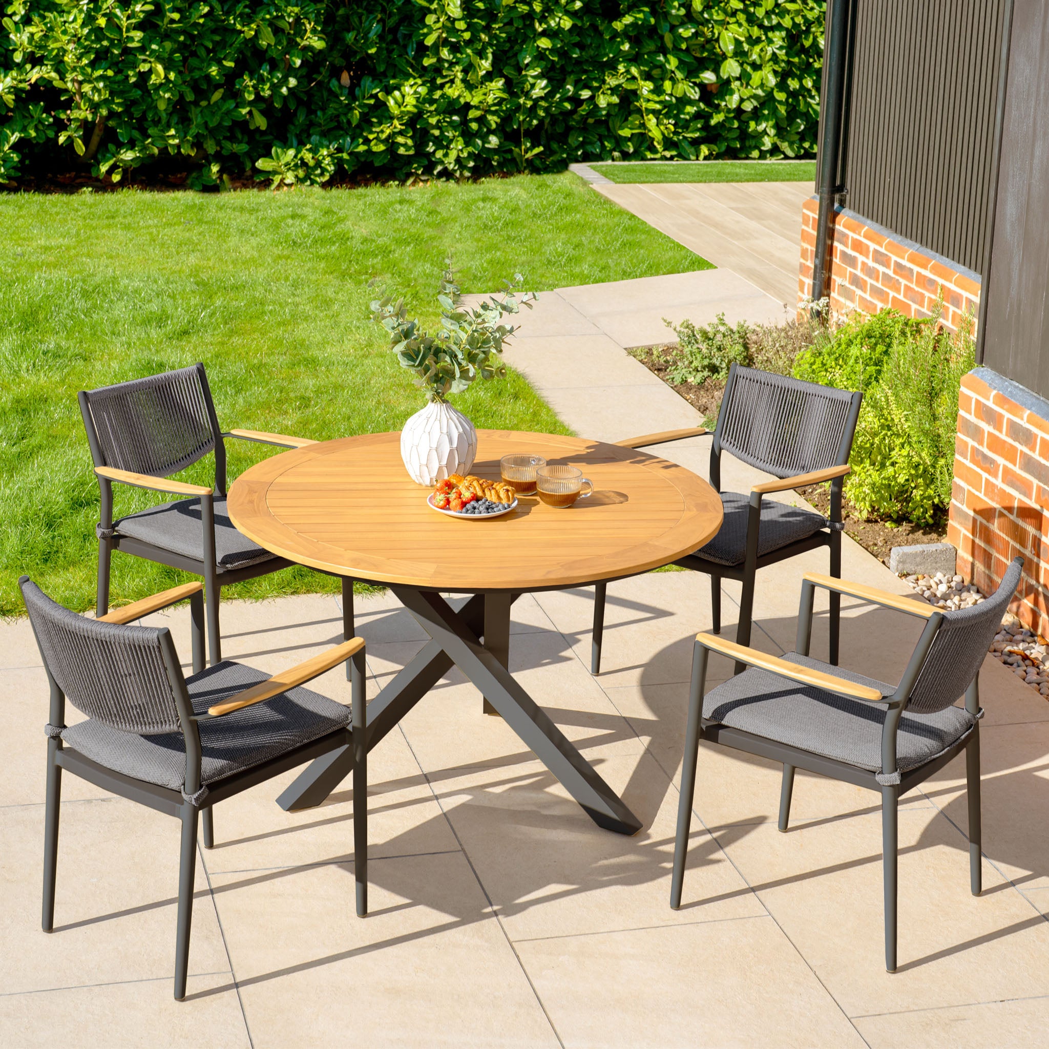 The Tellaro 4 Seat Round Dining Set with Teak Table in Charcoal sits elegantly on a tiled terrace, surrounded by lush greenery and a brick wall, offering a perfect outdoor retreat with its weatherproof rope chairs.