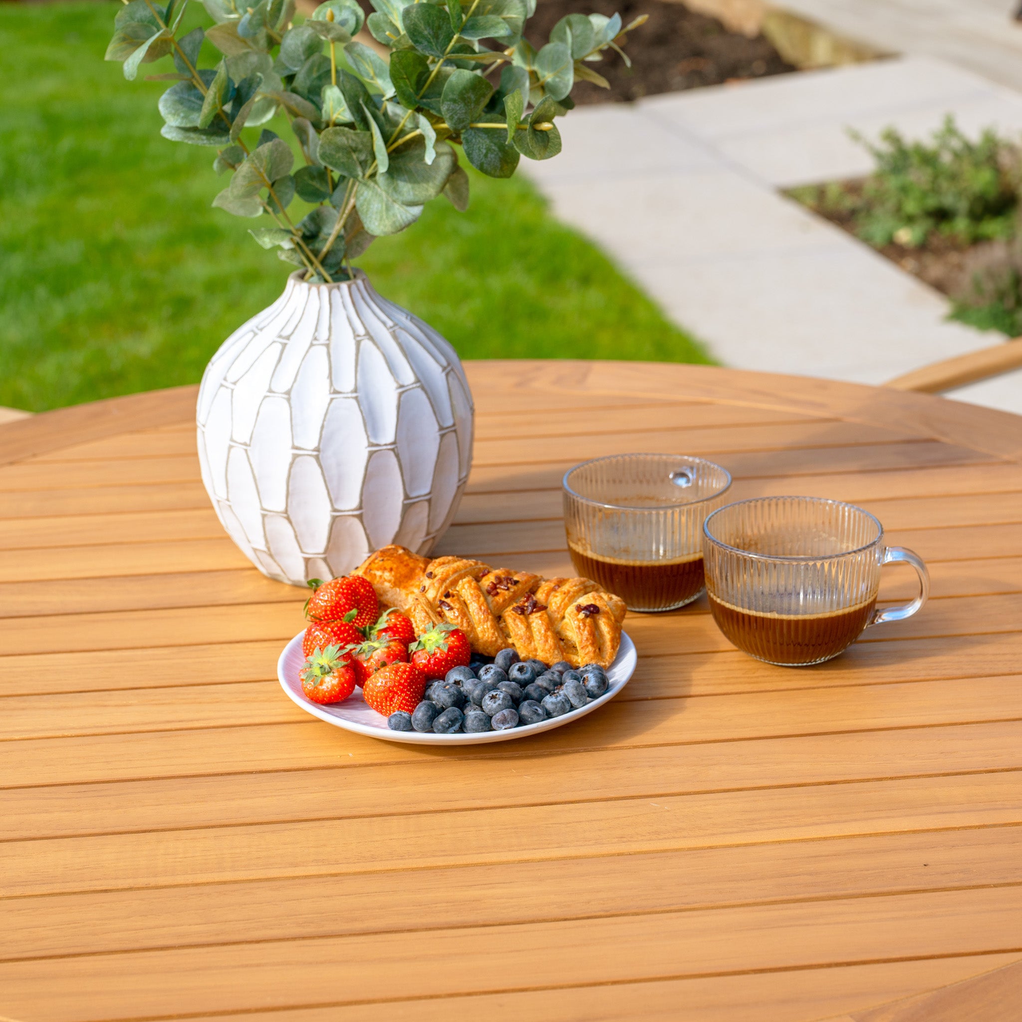 A plate of pastries, strawberries, and blueberries sits by two coffee cups on the Tellaro 4 Seat Round Dining Set with a teak table in charcoal. Weatherproof chairs enhance the outdoor setup, complemented by a vase of leaves for a natural touch.