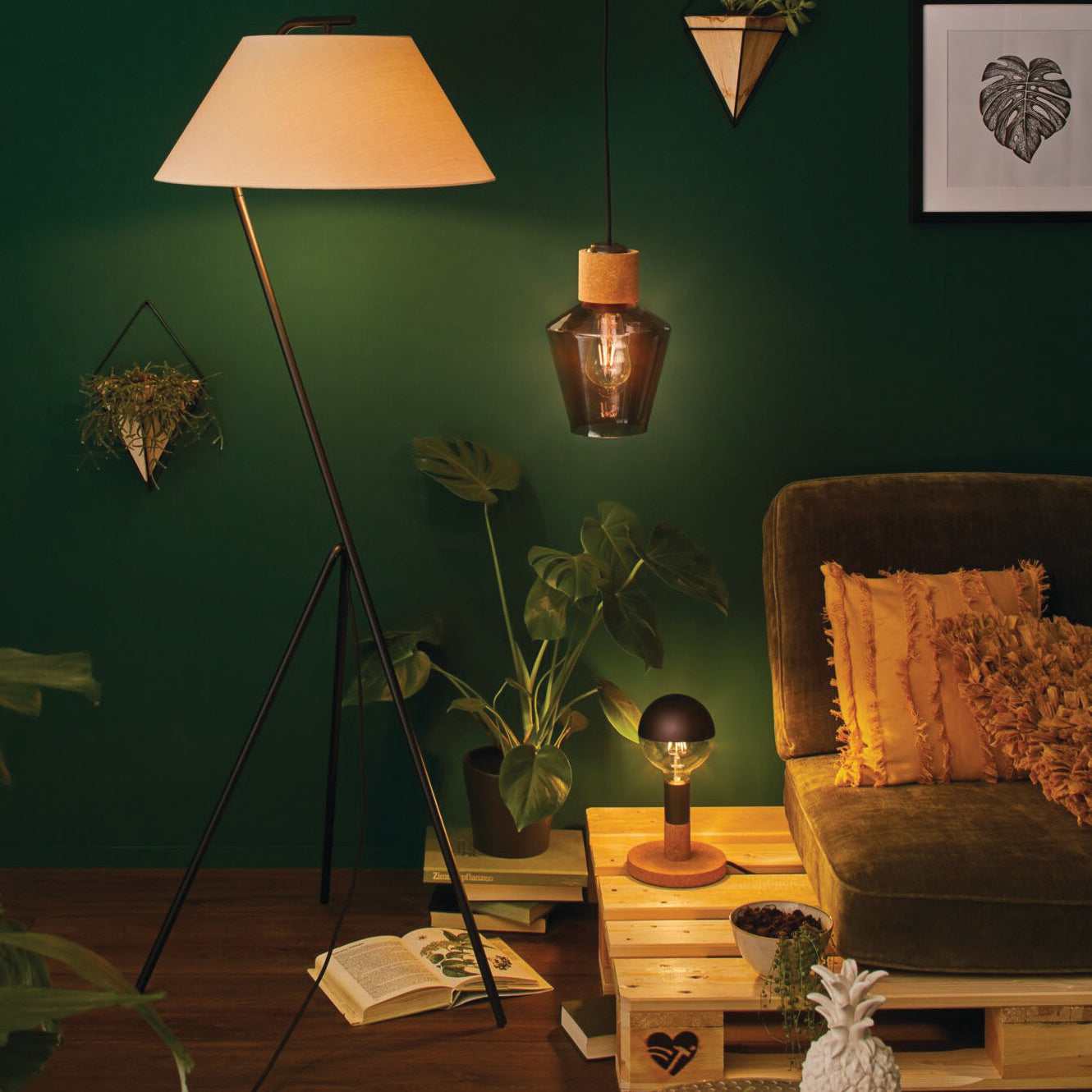 A cozy living room with green walls, various plants, a Standard LED Corn Bulb 6.5W E27 Gold Light on the floor, a retro style pendant luminaire hanging from the ceiling, and an orange pillow on a green sofa.