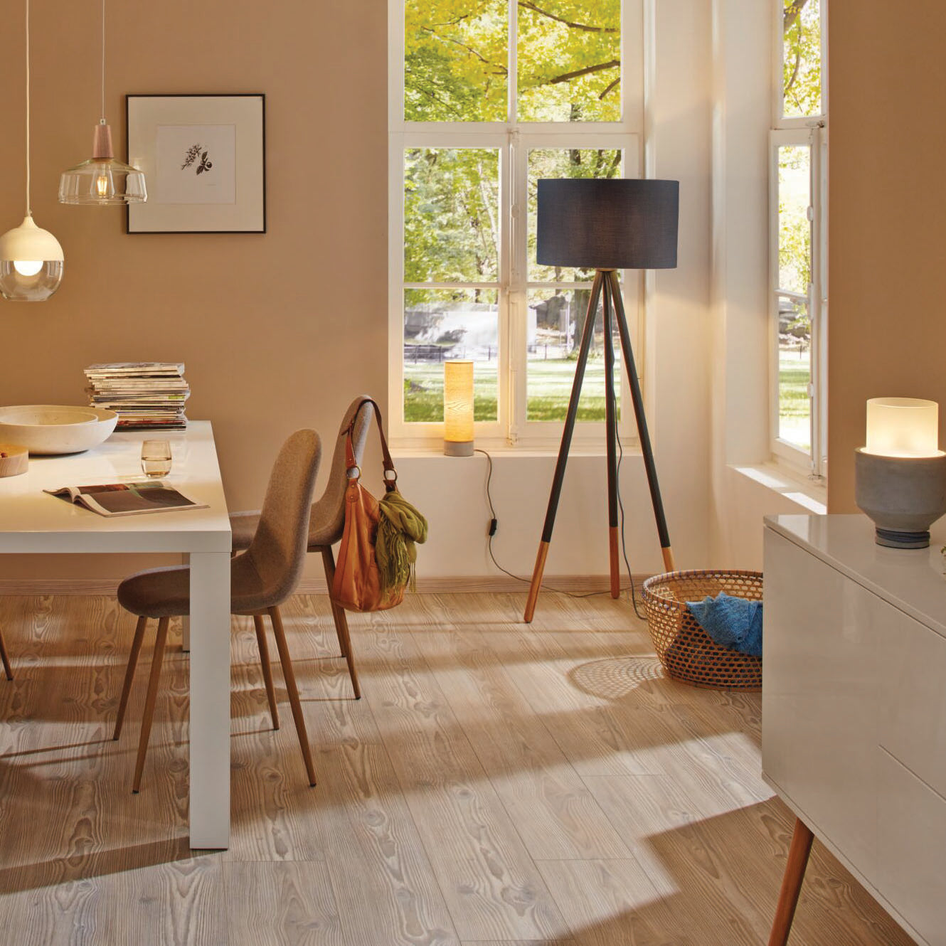 A cozy room featuring a white table and brown chairs is illuminated by a tall vintage lamp with the Paulmann - Standard LED Corn Bulb 6.5W E27 Gold Light. Large windows provide views of trees and ample sunlight, complemented by retro-style pendant luminaires that add a charming touch to the space.