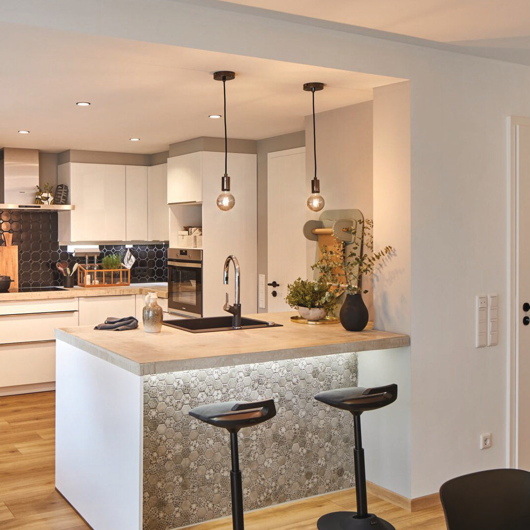 The modern kitchen features a wooden island with two stools and pendant lights showcasing the Paulmann Filament LED Globe Bulb 4W E27 in Gold Glass. Hexagonal tile accents on the wall contribute vintage charm, while the energy-efficient LED technology offers both style and sustainability.