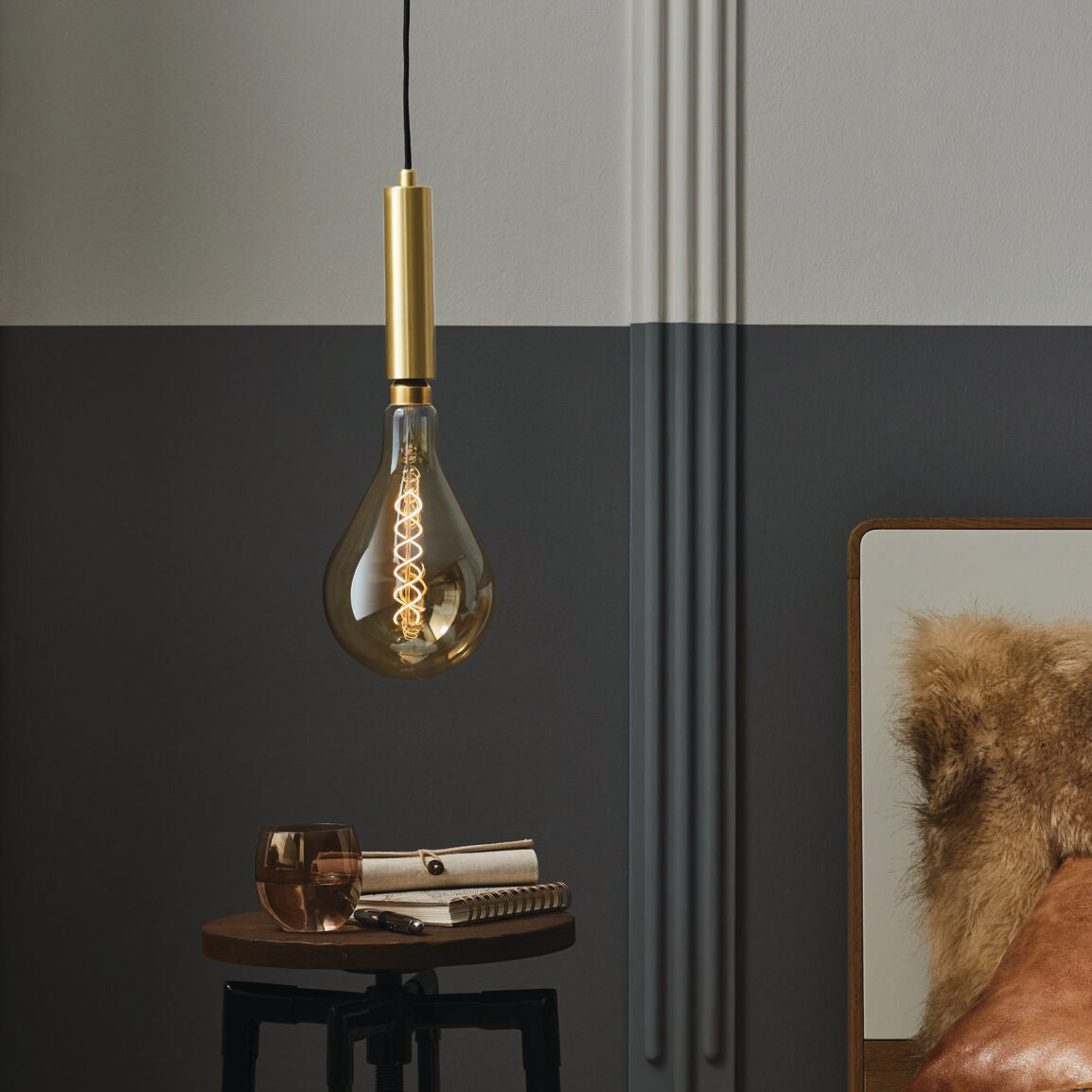 A Paulmann Filament LED Big Drop Bulb 7W E27 in Gold light casts a warm glow over the vintage-style interiors, illuminating a small table with a notebook and glass, while a cozy fur blanket invites comfort on the nearby bed.