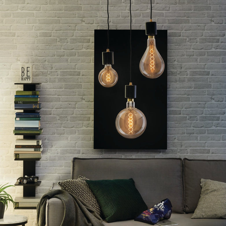 In a modern living room with vintage-style interiors, you'll find hanging Paulmann Filament LED Globe Bulbs in 4W E27 Smoke Glass, a gray sofa, stacked books, and a "Be Happy" sign on the wall. The energy-efficient LED technology enriches the ambiance while remaining eco-friendly.