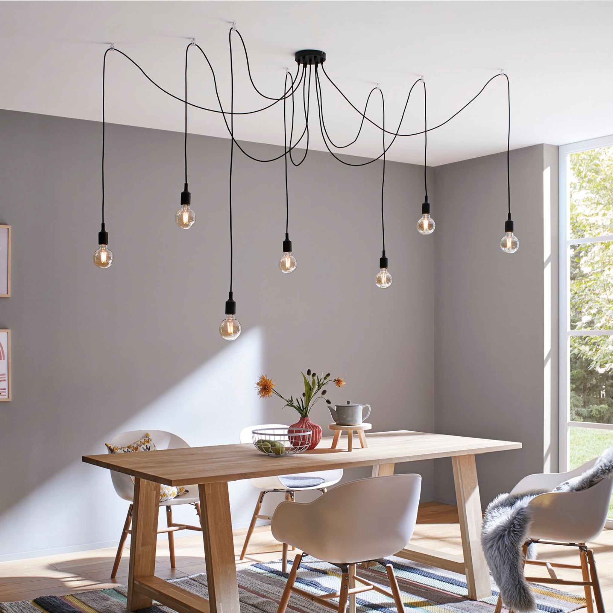 A contemporary dining room showcasing decorative lighting with Edison bulbs includes the Paulmann Neordic Ketil 420W 7 Lamp Dimmable Pendant Light in Black above a wooden table, complemented by white chairs and a large window that invites sunlight.