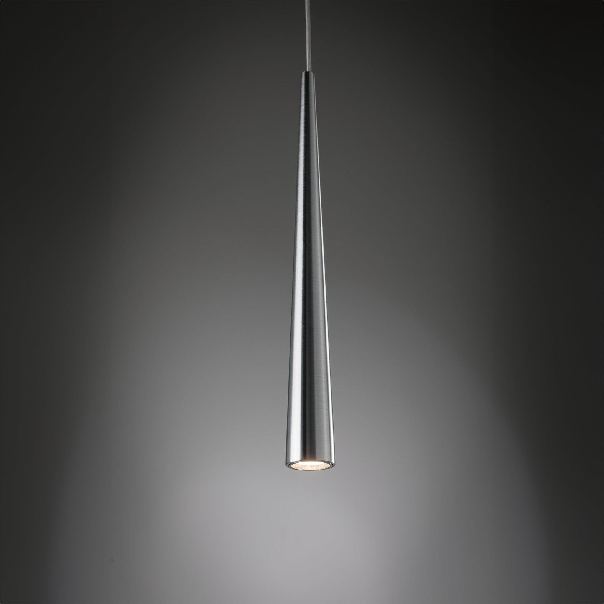 A sleek Gutta 8.5W LED Dimmable 1 Lamp Pendant Light in Brushed Aluminium with an adjustable fabric cable hangs against a dark background, emitting a soft, focused lighting effect at the bottom.