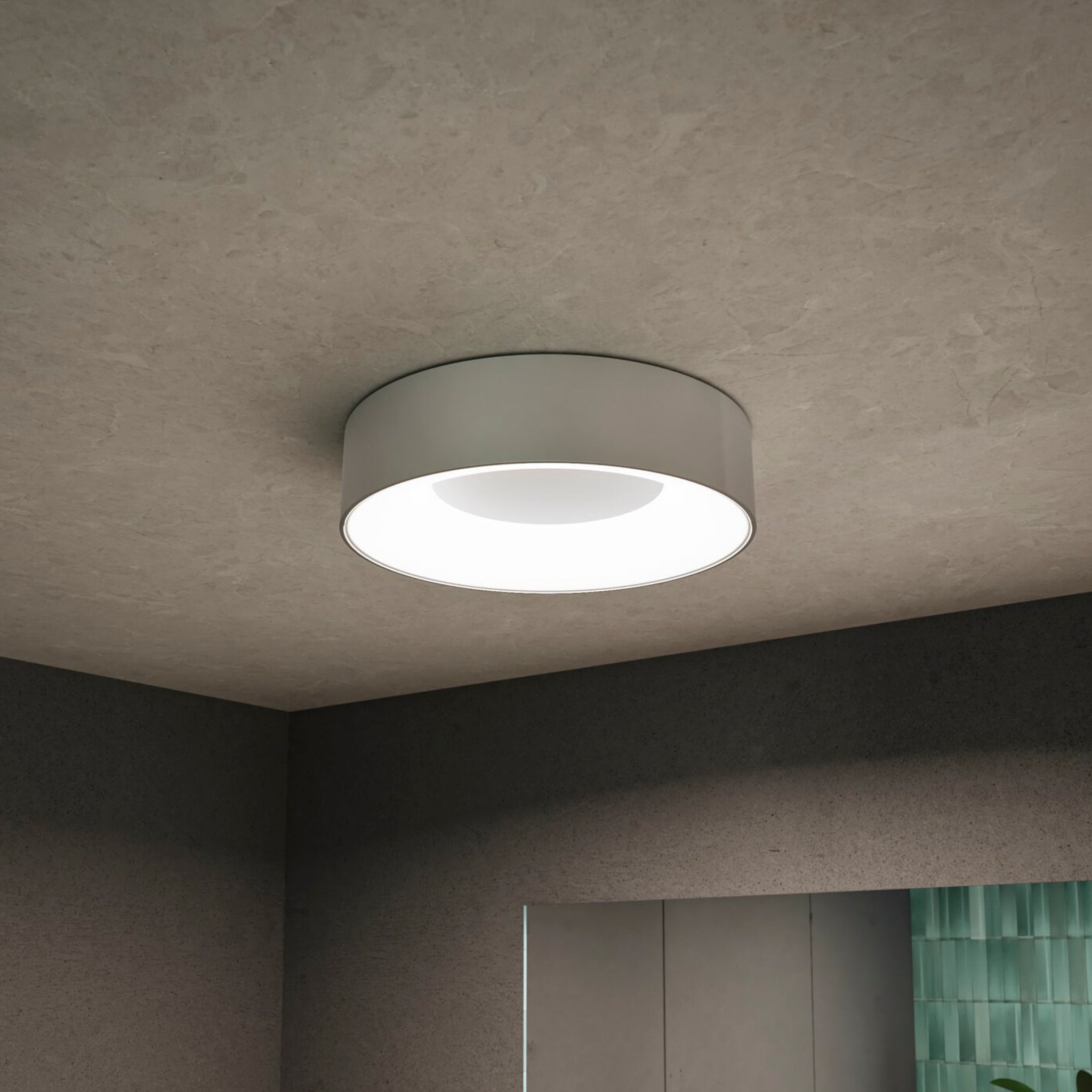 The Casca 18W LED Switch 1500lm Ceiling Light in Matt Aluminium features a modern design, ideal for illuminating rooms with textured walls. It offers adjustable light color settings for a unique lighting effect, while glimpses of green-tiled areas enhance the overall aesthetic.