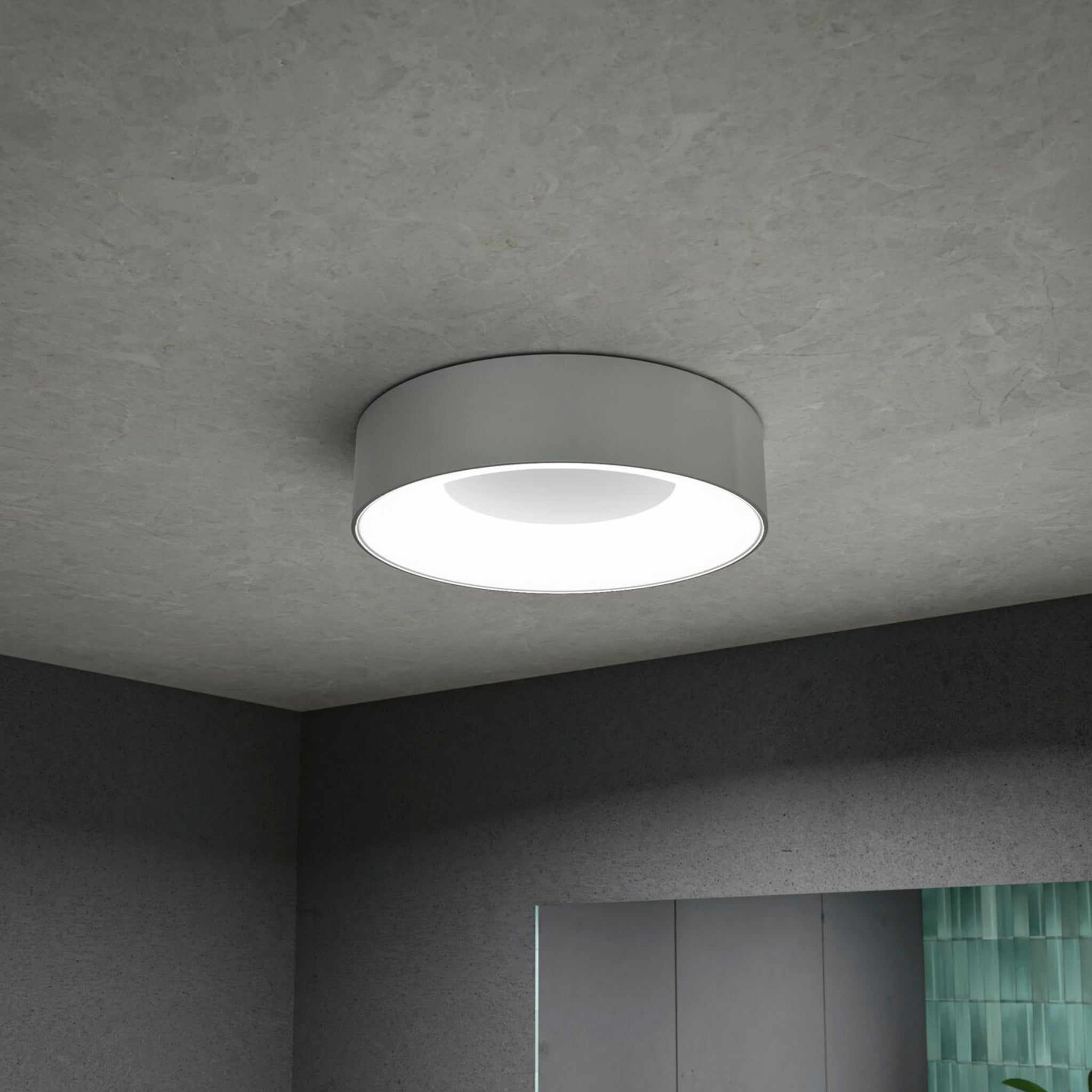 The Casca 18W LED Switch 1500lm Ceiling Light in Matt Aluminium features a round, modern design with a flat, circular profile, perfectly illuminating a minimalist grey room. This fixture offers a special lighting effect that allows you to adjust the light color to suit your mood or setting.