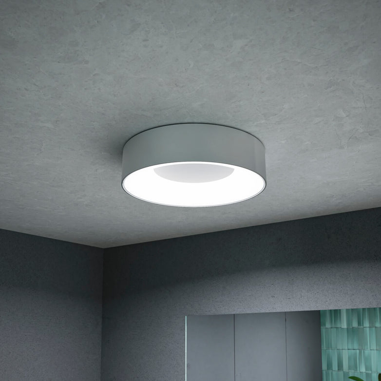 A modern Casca 18W LED Switch 1500lm Ceiling Light in Matt Aluminium with a sleek design illuminates a minimalist room with gray walls and ceiling, offering the special lighting effect to adjust light color.