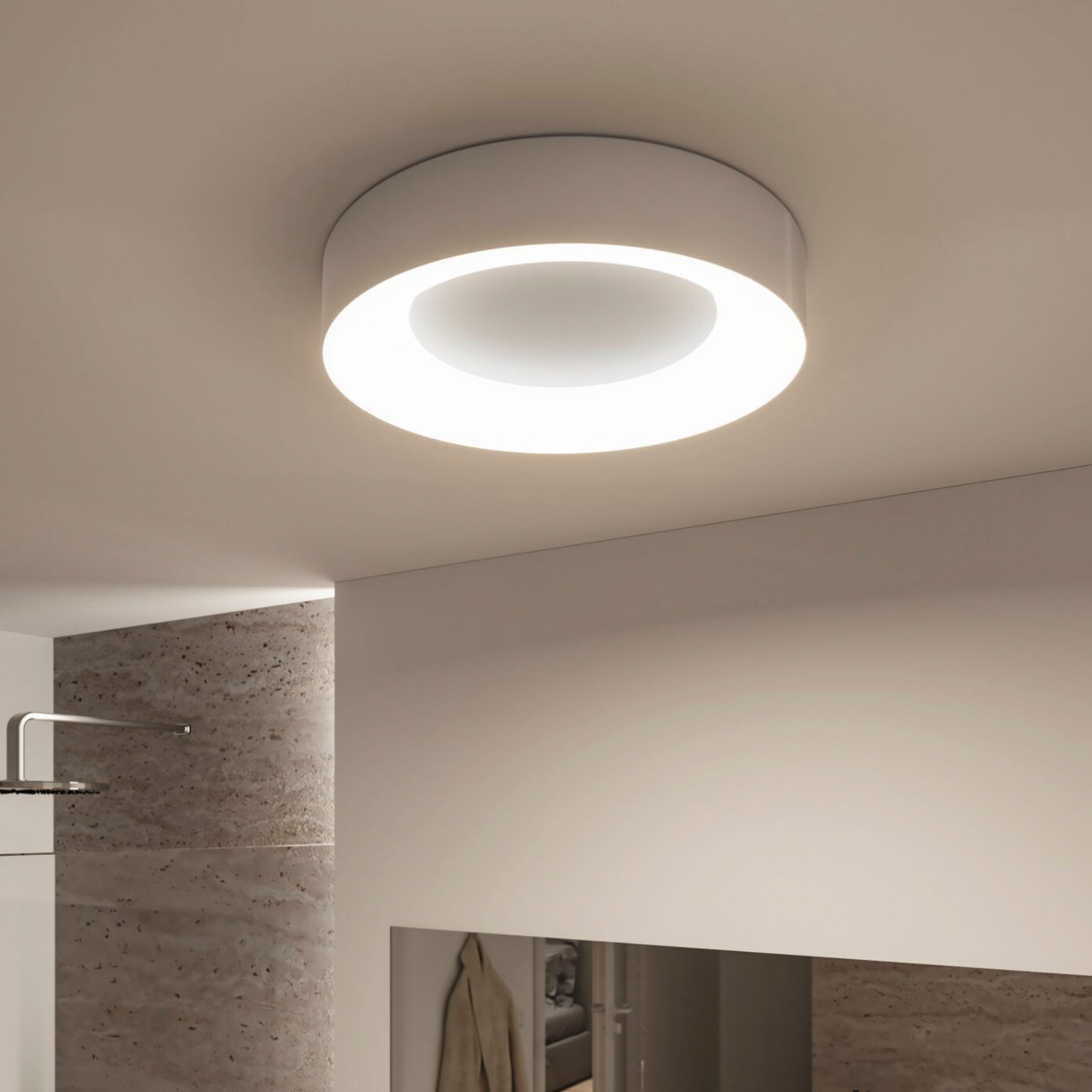 A modern Casca 25W LED Switch 2100lm Ceiling Light in Matt Aluminium emits white light in a minimalist room with beige walls and a mirror, offering a special lighting effect that adjusts the light color to create the perfect ambiance.