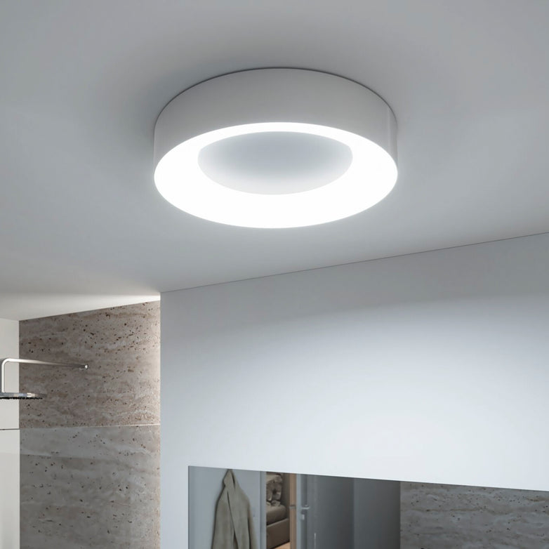 The **Casca 25W LED Switch 2100lm Ceiling Light in Matt Aluminium**, with its modern circular design, illuminates a minimalist room with light-colored walls and a mirror, offering a special lighting effect that adjusts the light color.