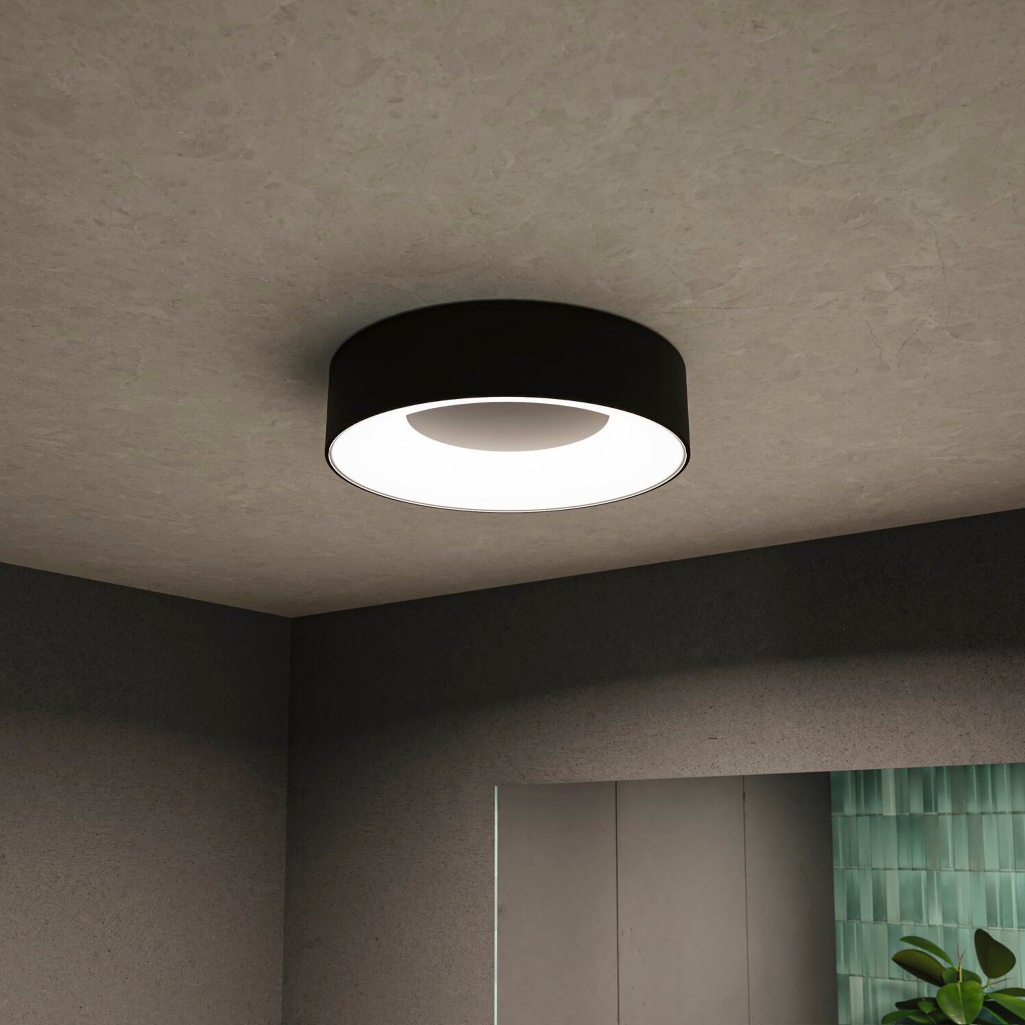 A black matt Casca 18W LED Switch 1500lm Ceiling Light illuminates a modern room with a special lighting effect, highlighting a partial view of a mirror and a plant.