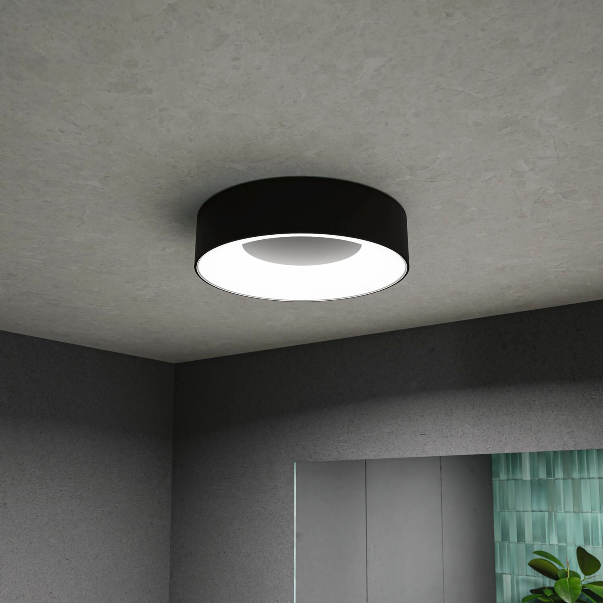 The Casca 18W LED Switch 1500lm Ceiling Light in Black Matt, a sleek and modern circular fixture, illuminates a contemporary room with a tiled wall and partial plant view, creating a special lighting effect.