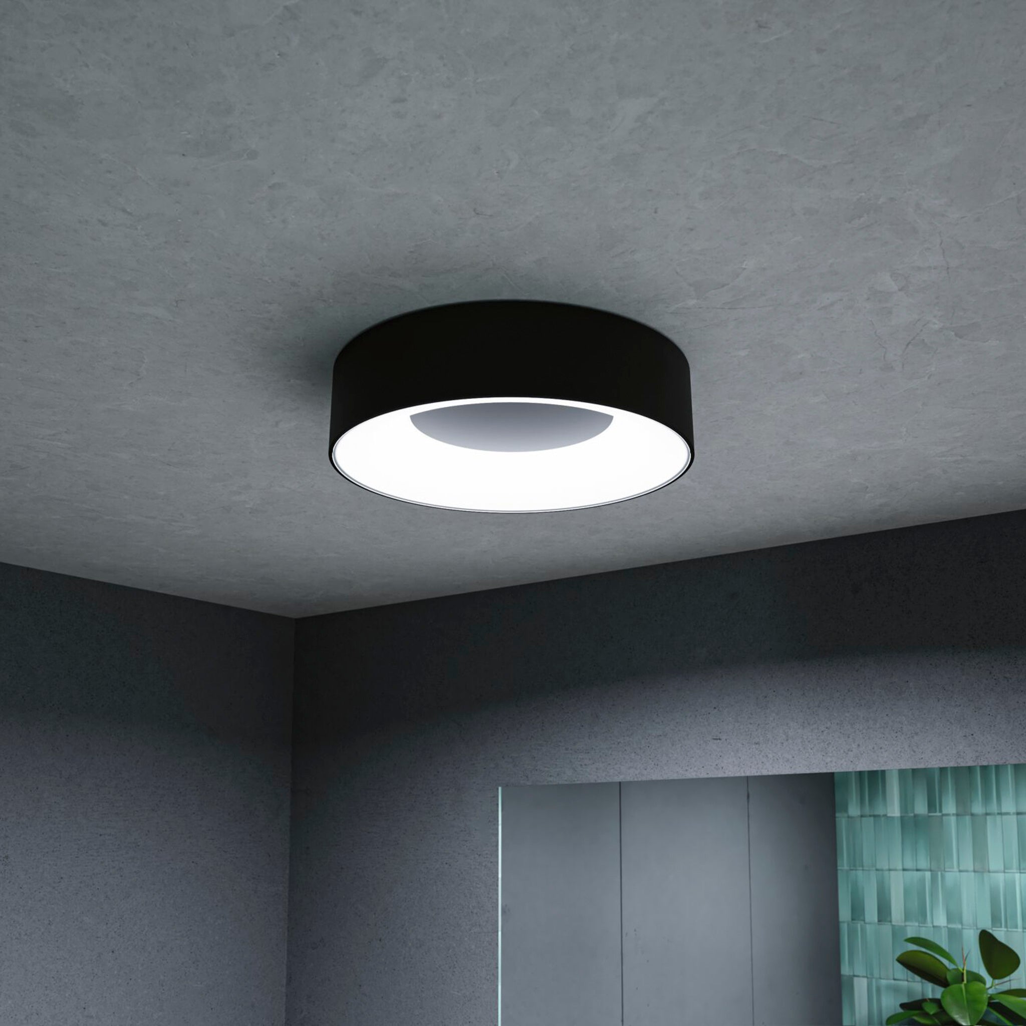 The Casca 18W LED Switch 1500lm Ceiling Light in Black Matt is a contemporary circular fixture, elegantly mounted on a gray ceiling adorned with green accents. Its distinctive lighting effect not only enhances the light's color but also cultivates an inviting atmosphere, making any room feel exceptionally welcoming.