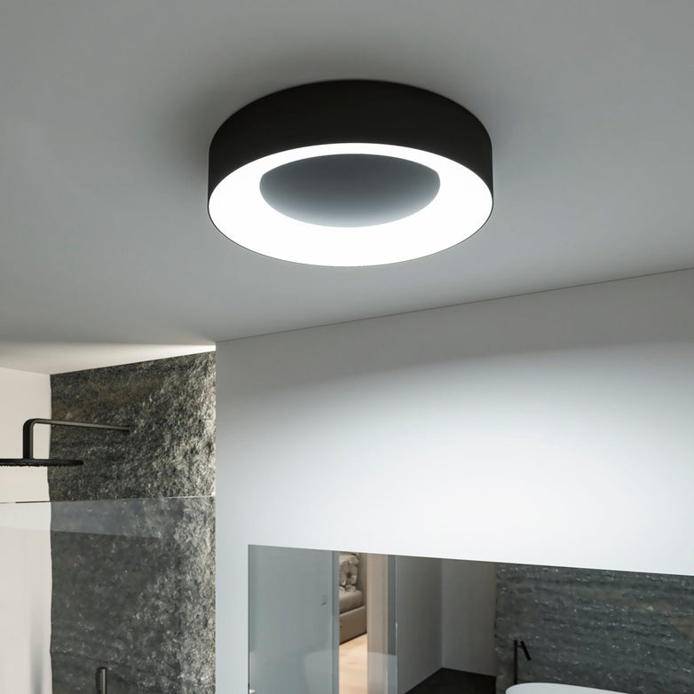 The Casca 25W LED Switch 2100lm Ceiling Light in Black Matt illuminates a minimalistic room with textured walls, offering an adjustable light color for the perfect lighting effect.