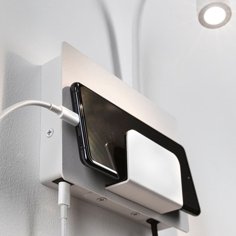 A Halina 3.5W LED Wall Light with USB Charging Port in White Matt, featuring a flexible luminaire arm and connected USB cable, is holding a smartphone.