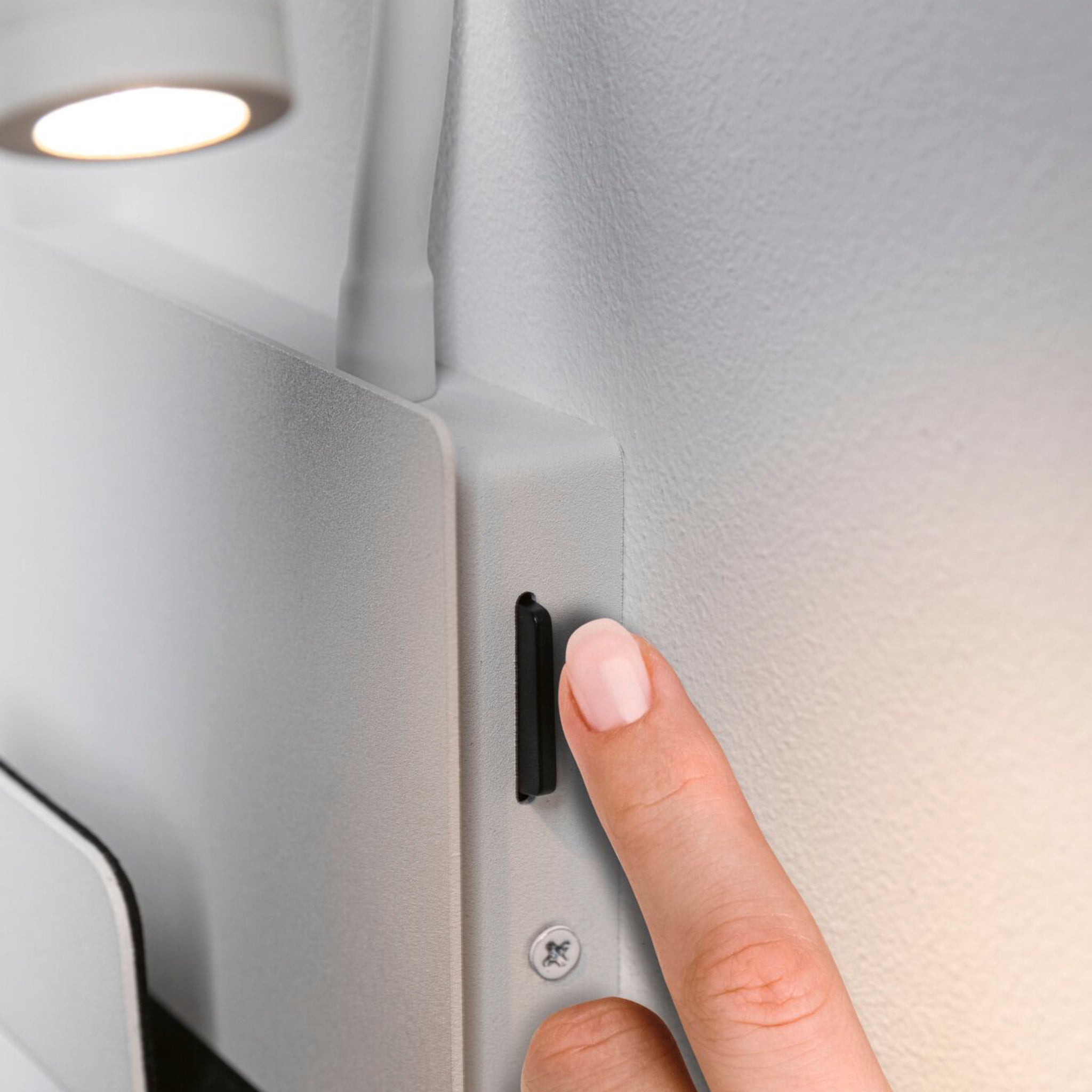 A hand with a pink manicure presses a black button on a Halina 3.5W LED Wall Light with USB Charging Port in White Matt, mounted on the wall under its small, lit lamp with a USB Type-C charging socket.