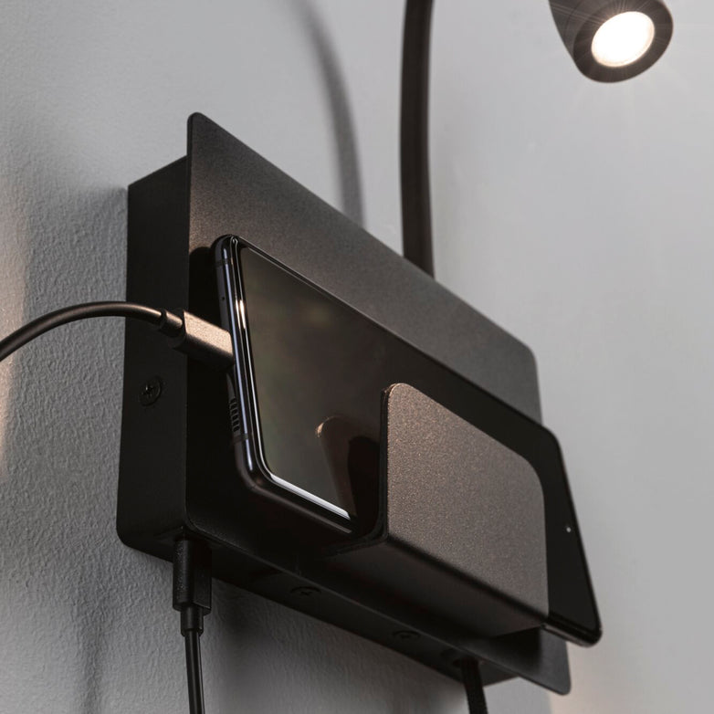 Phone charging on a wall-mounted wireless charger under the small, adjustable Halina 3.5W LED Wall Light with USB Charging Port in Black Matt, featuring a flexible arm.