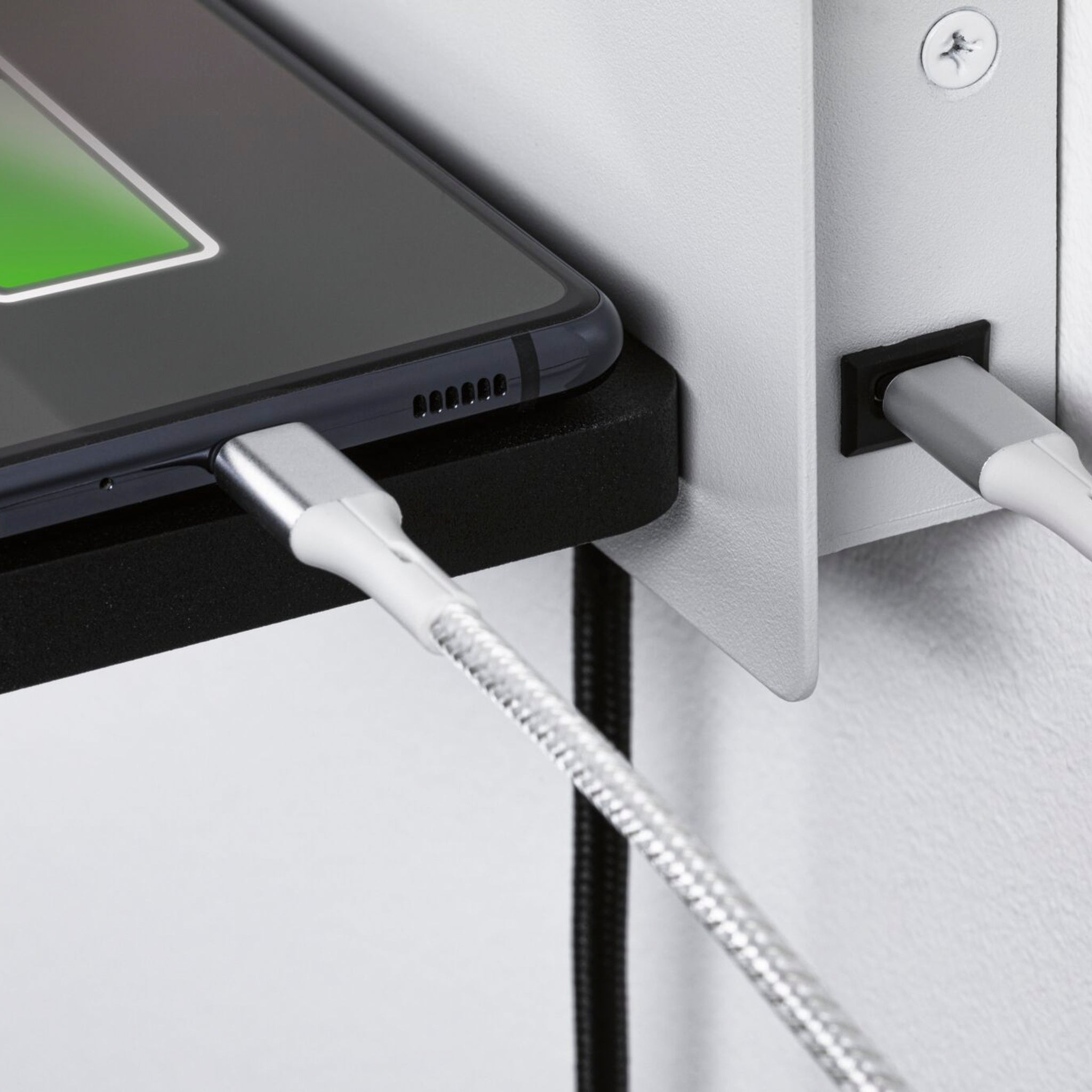 A close-up view of a smartphone with a USB Type-C charging port, connected via a silver cable to the wall-mounted USB port of the Paulmann - Serra 5.5W/2.6W LED Right Hand Wall Light with 3-Step Dim in White & Black, which features a sleek design suitable for reading.