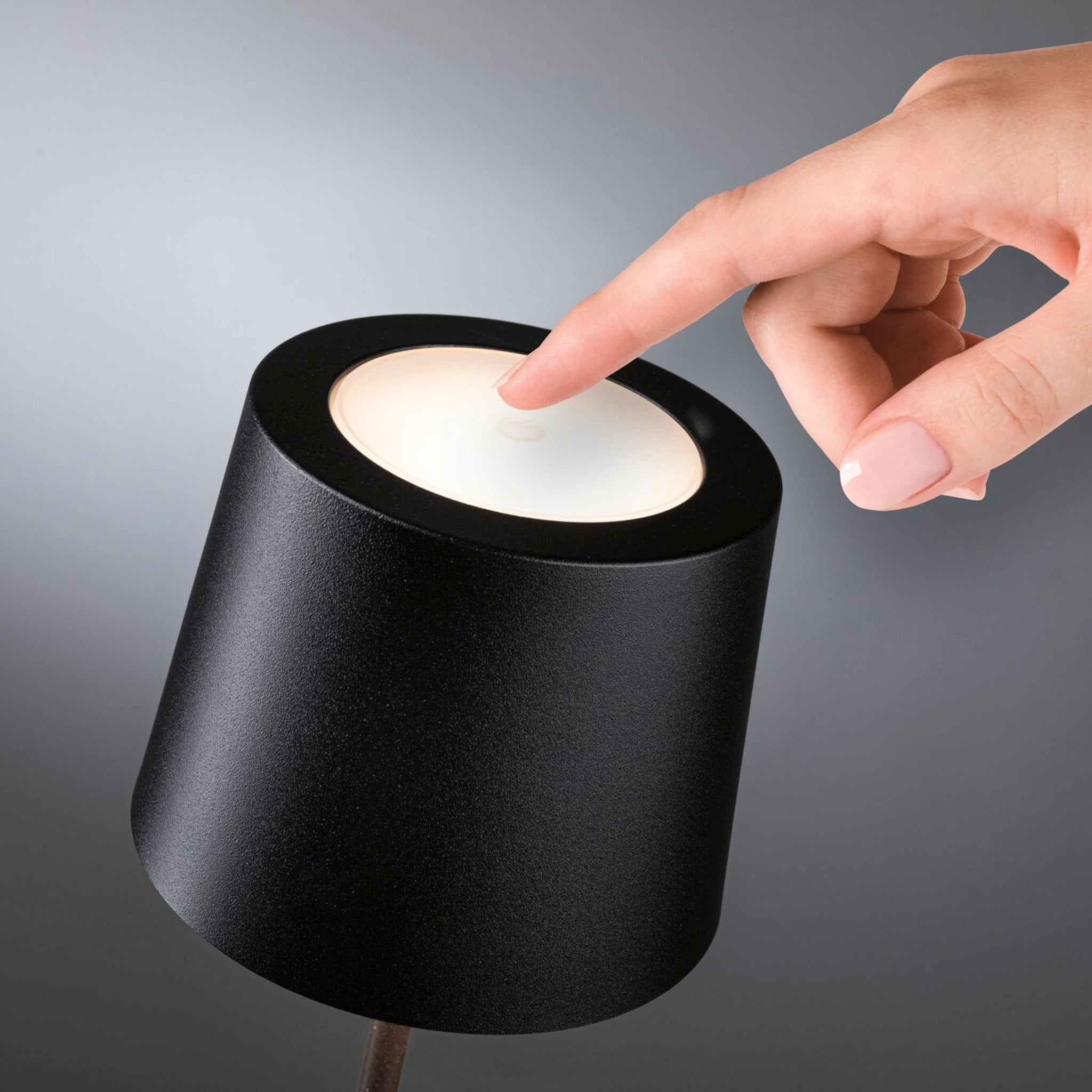 A hand is about to touch the top of a Gilo 2.8W LED Mobile Dimmable Floor Lamp in Black Matt, activating its continuous touch dimming function for a warm white lighting mood. This sleek, portable battery-powered LED table luminaire combines convenience and style seamlessly.