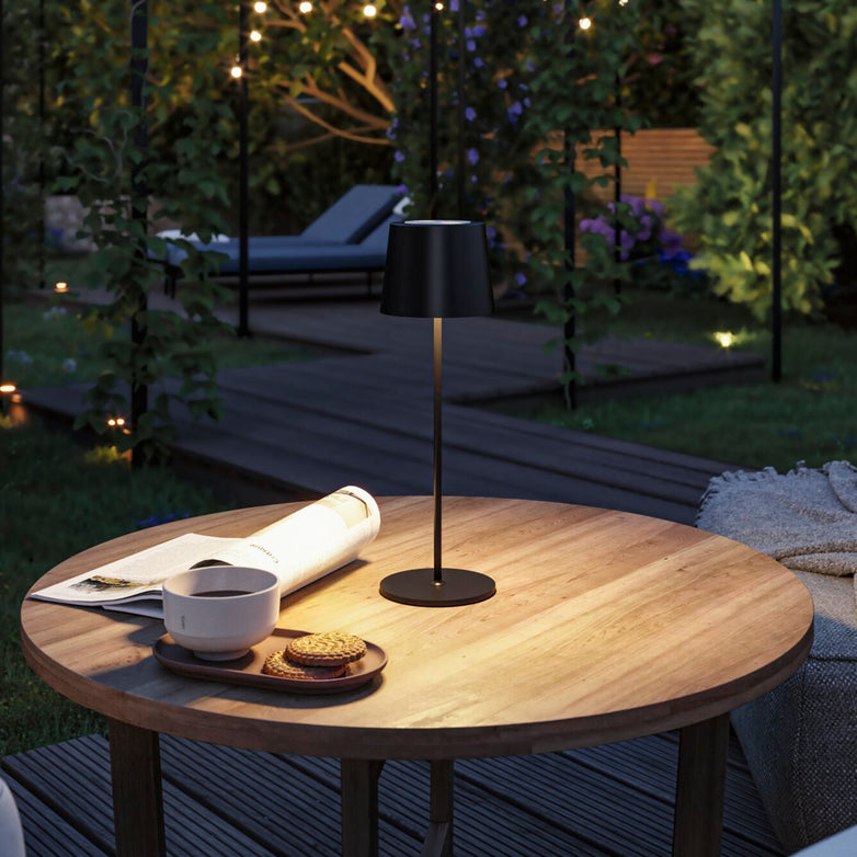 A cozy outdoor evening setup features a wooden table adorned with snacks, a cup, and a rolled magazine, all gently illuminated by the Gilo 2.8W LED Mobile Dimmable Floor Lamp in Black Matt. The warm white lighting mood and continuous touch dimming function add to the ambiance.