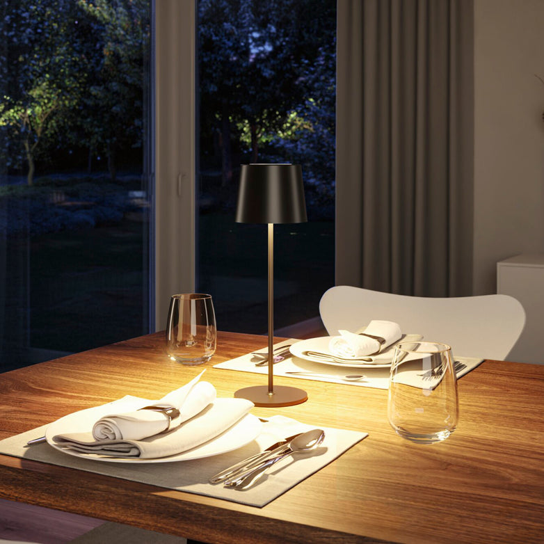 A dining table set for two featuring the Gilo 2.8W LED Mobile Dimmable Floor Lamp in Black Matt at its center, providing a warm white lighting mood. The sleek, portable lamp with a continuous touch dimming function is placed beside a window overlooking a night garden.