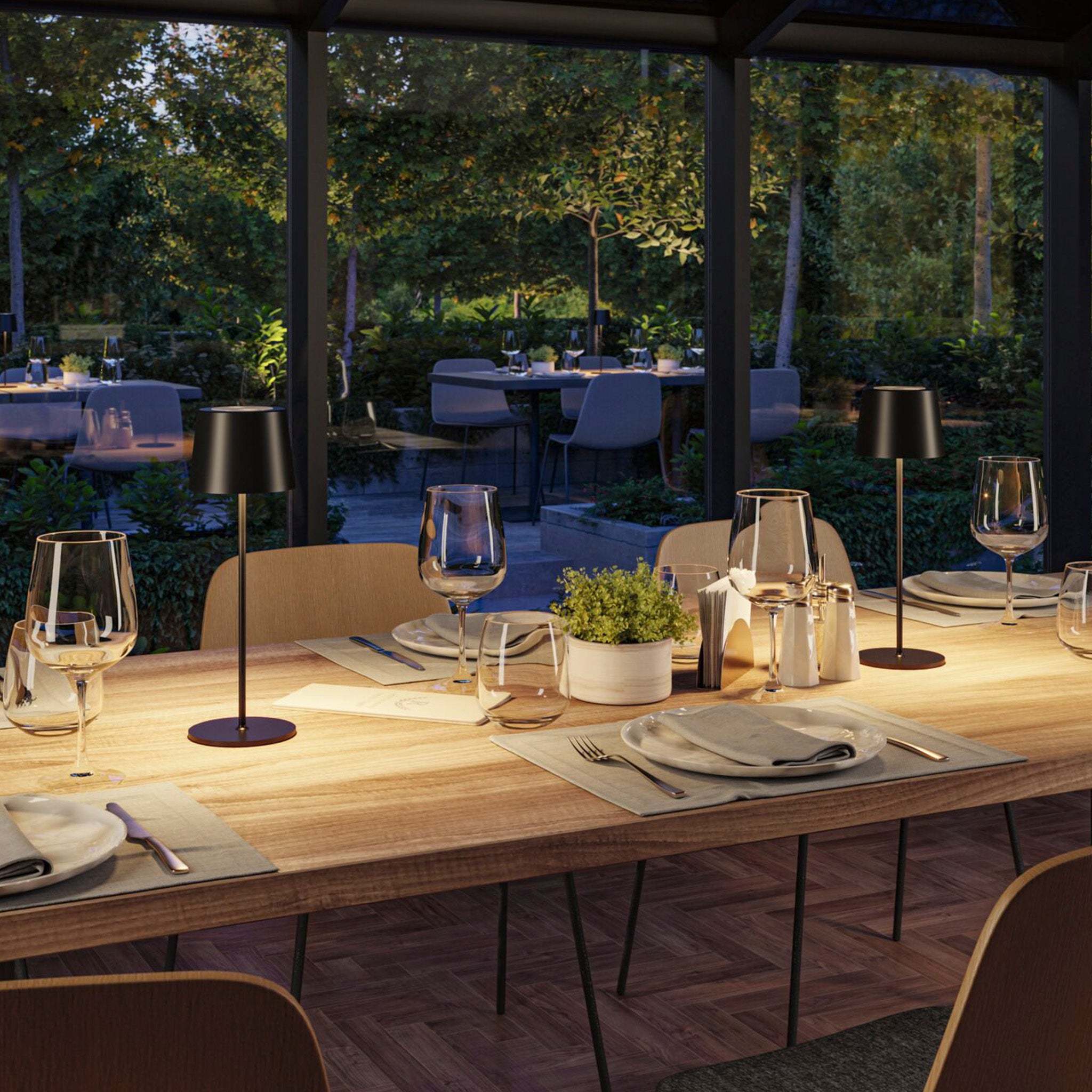 A modern restaurant dining table set with plates, wine glasses, and small lamps featuring the Gilo 2.8W LED Mobile Dimmable Floor Lamp in Black Matt. The warm white lighting mood and continuous touch dimming function create an inviting ambiance, with a beautiful garden view through glass walls.