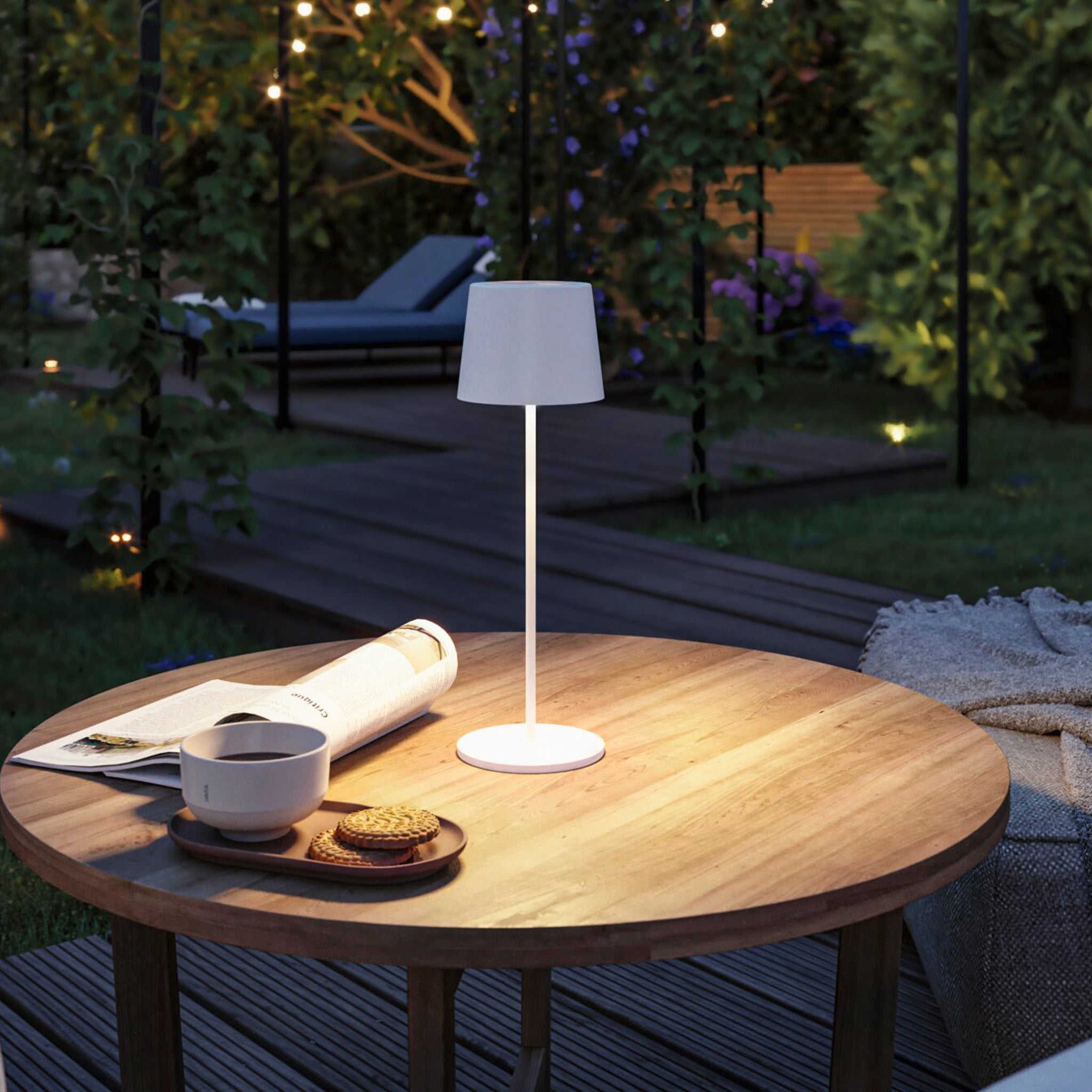 Gilo 2.8W LED Mobile Dimmable Floor Lamp in White Matt lights up the outdoor table, complemented by tea and cookies under the night sky. The background features garden lights, lush greenery, and a cozy lounge chair. The USB-C touch dimming feature adjusts to create the perfect ambiance for any setting.