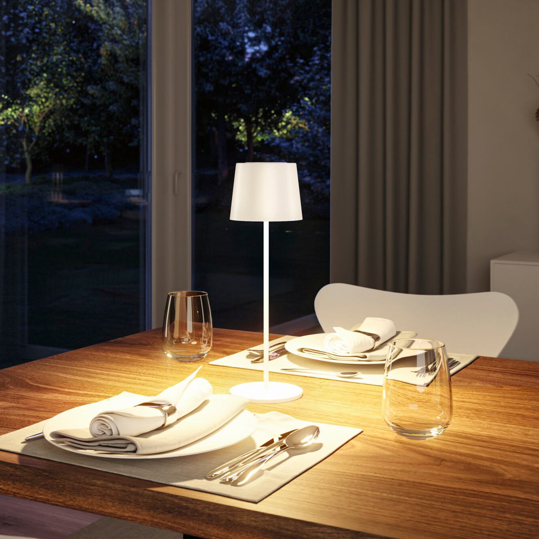 A dining table set for two with plates, cutlery, napkins, and glasses, softly illuminated by the Gilo 2.8W LED Mobile Dimmable Floor Lamp in White Matt featuring USB-C touch dimming in the dimly lit room.