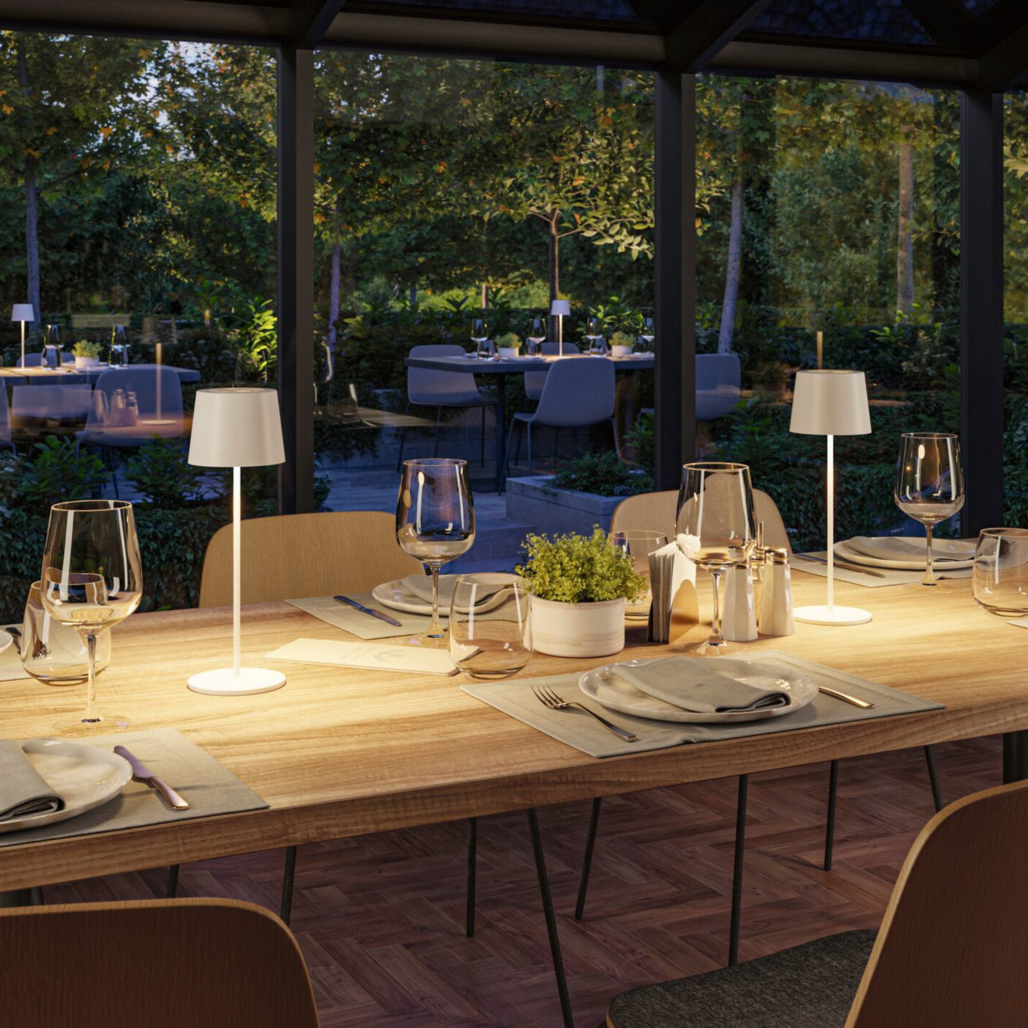 Elegant restaurant setting with an outdoor garden view, featuring a wooden table, modern decor, and soft ambient lighting enhanced by the Gilo 2.8W LED Mobile Dimmable Floor Lamp in White Matt.