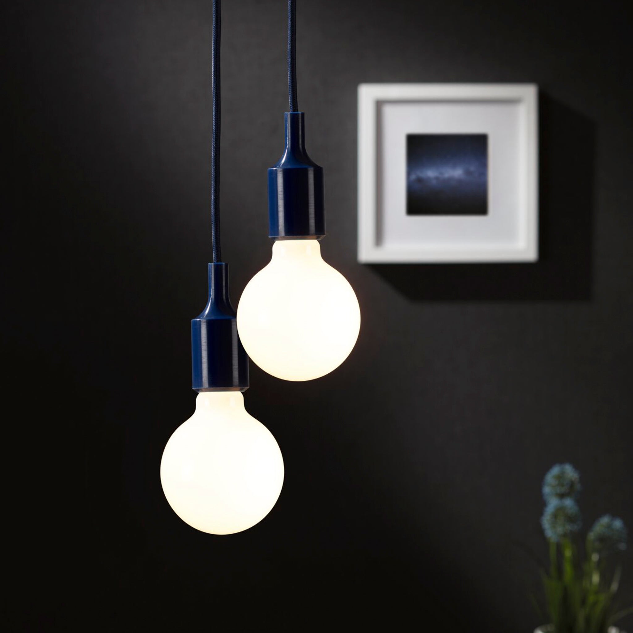 A pair of Paulmann Neordic Ketil 60W dimmable pendant lights, featuring night blue silicone holders, cast a warm glow against a dark wall adorned with a framed picture.