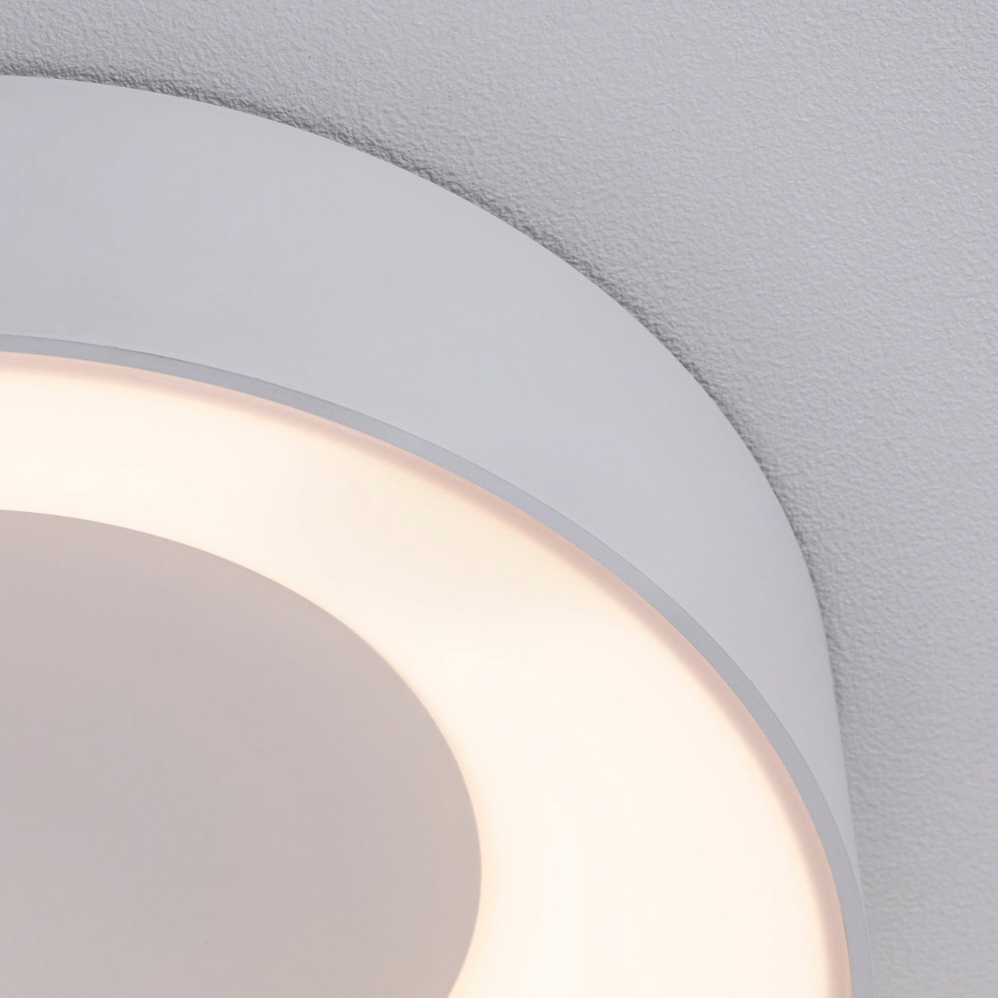 Close-up of the Casca 18W LED Switch 1500lm Ceiling Light in White, a modern, circular luminaire with adjustable light color, casting a soft, warm glow against a textured white ceiling.