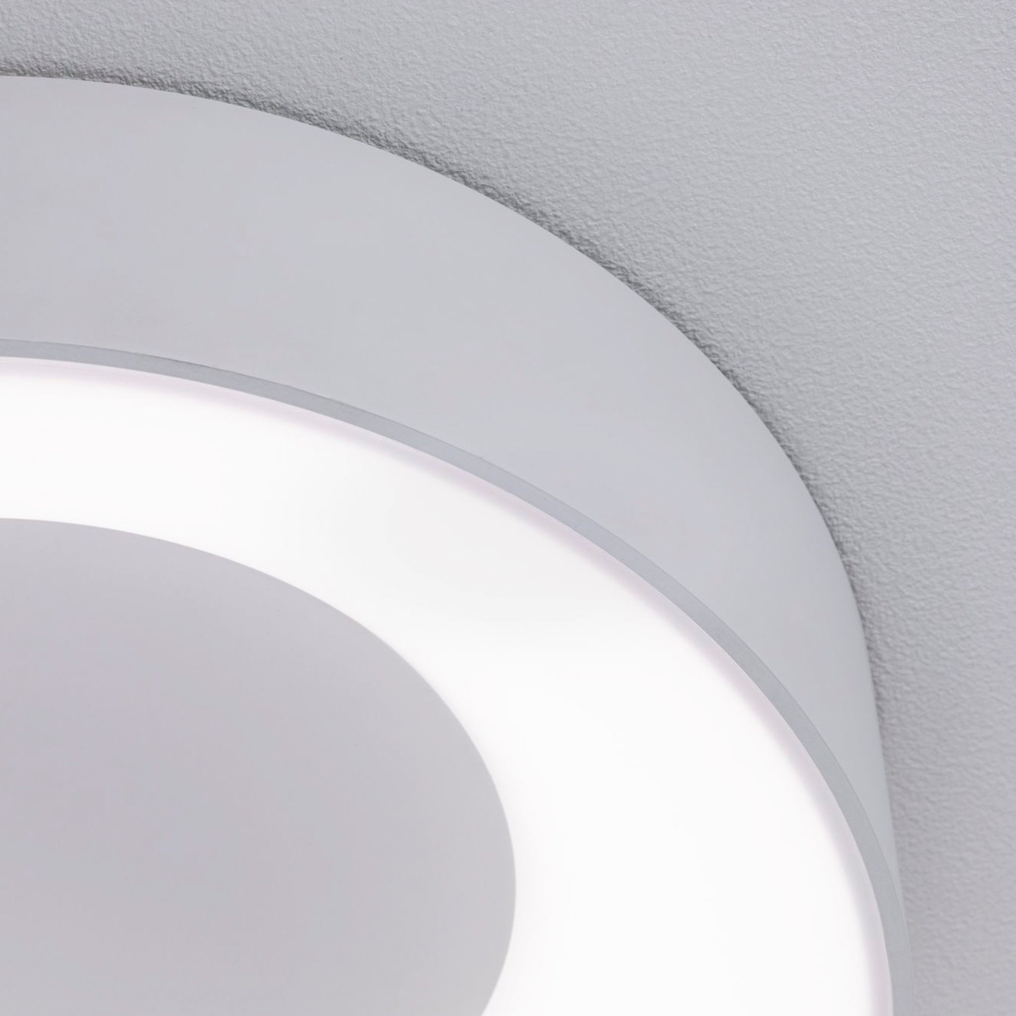 Close-up of the Casca 18W LED Switch 1500lm Ceiling Light in White, featuring a modern circular design with a white ring emitting soft light against a gray ceiling. The luminaire offers adjustable light color to suit any mood.