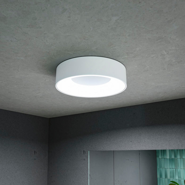 The Casca 18W LED Switch 1500lm Ceiling Light in White perfectly complements the modern, minimally decorated room with its round design, seamlessly blending into the grey ceiling and wall while offering an adjustable light color for the ideal ambiance.