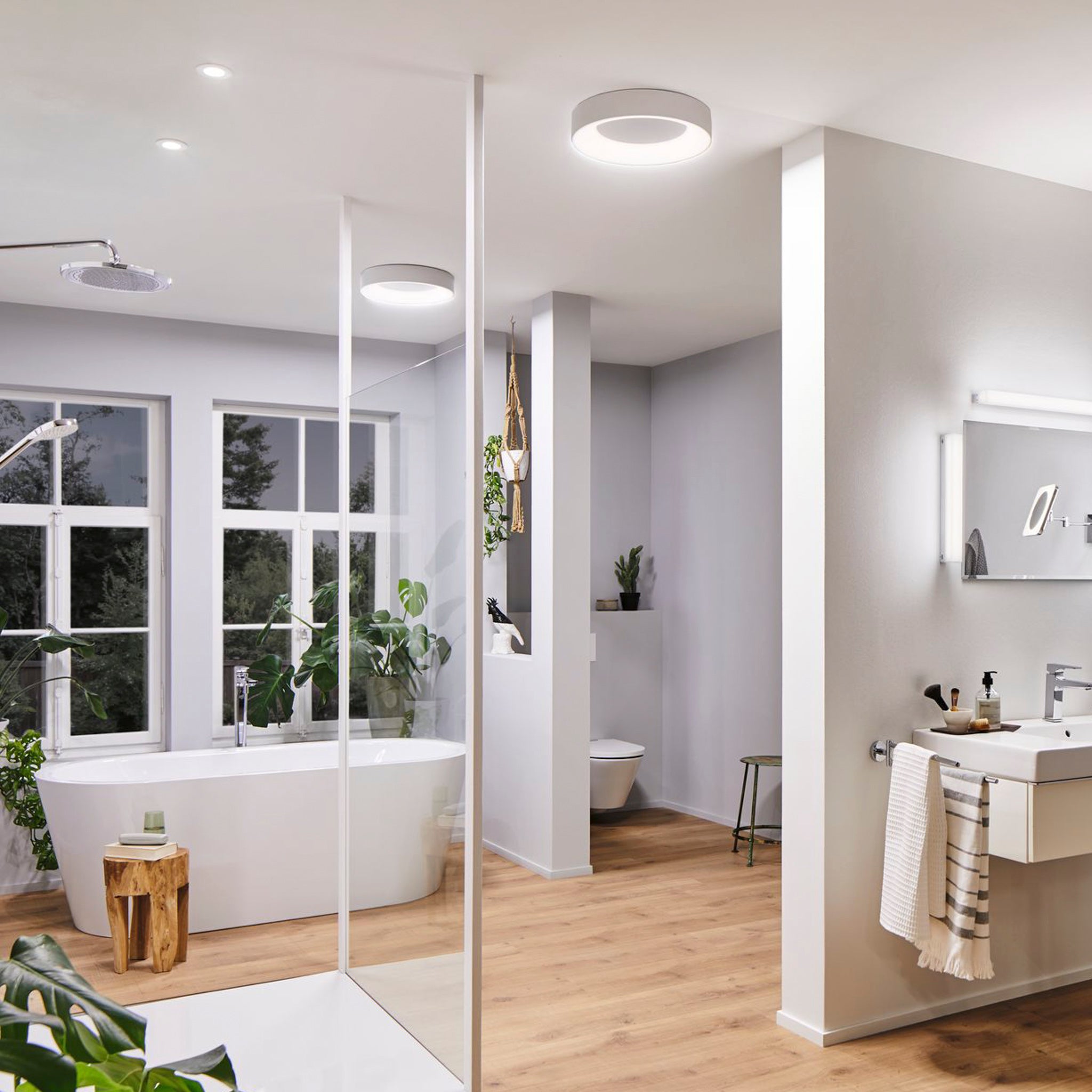 Modern bathroom design featuring a bathtub, shower, and vanity with a mirror, surrounded by green plants and natural light. An elegant Casca 25W LED Switch 2100lm Ceiling Light in White enhances the space, offering adjustable light color to create the perfect lighting effect for any mood.