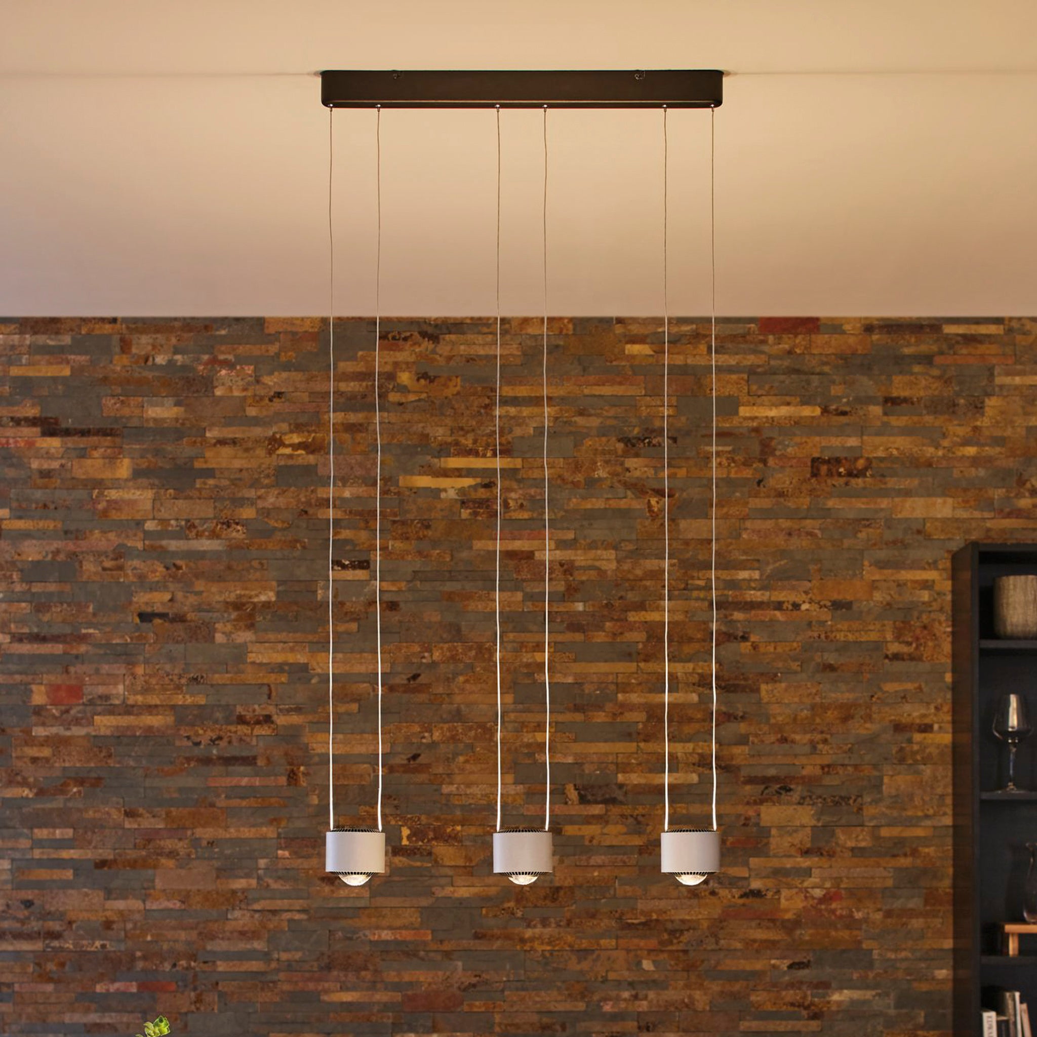 The Aldan 28W LED Dimmable 3 Lamp Pendant Light in Brushed Aluminium, featuring adjustable pendant lengths, hangs against a textured brick wall in a room defined by its contemporary and elegant design.