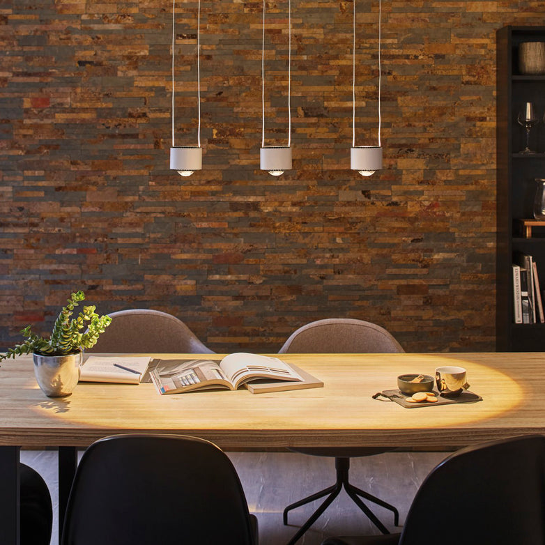 Modern dining room with wooden table, books, coffee cups, and Aldan 28W LED Dimmable 3 Lamp Pendant Light in Brushed Aluminium featuring adjustable lengths. Brick wall in the background.