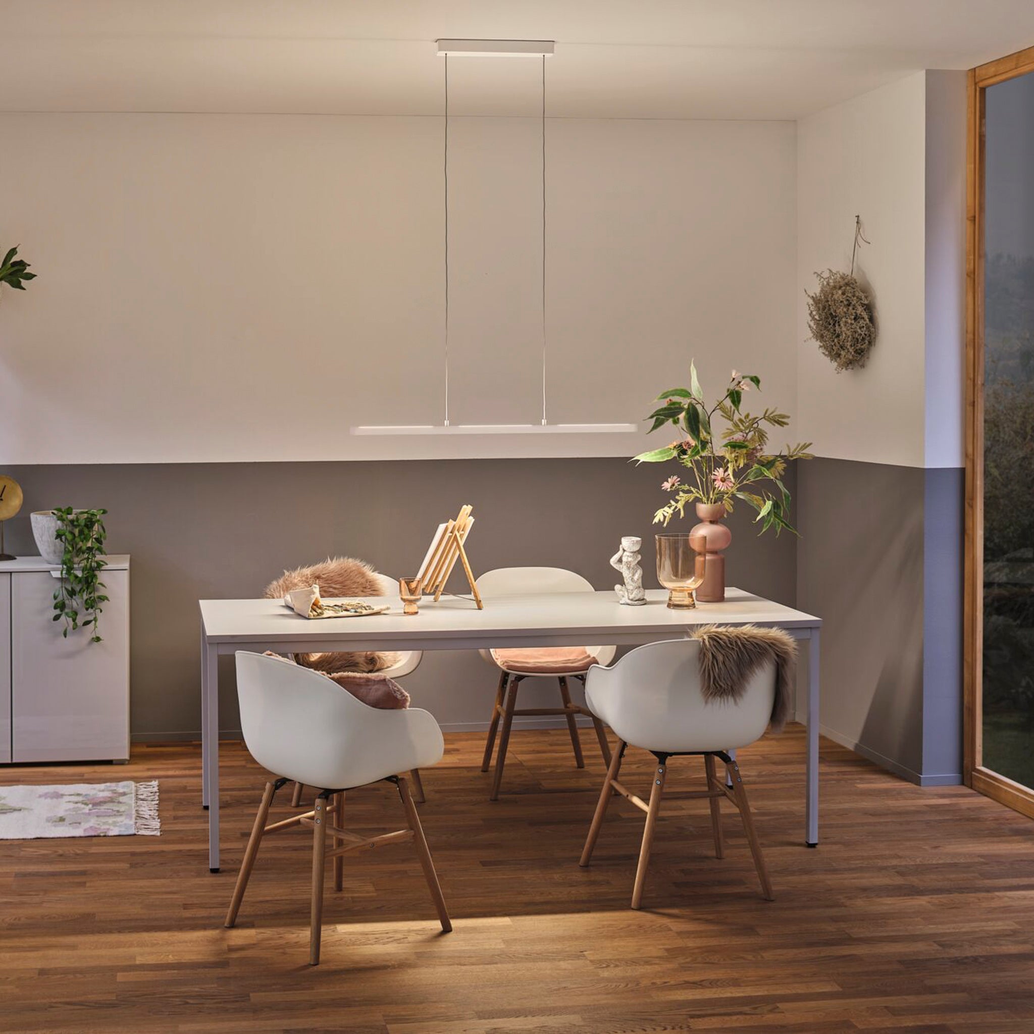 A modern dining room features a white table and four chairs, illuminated by the Paulmann Lento 37W LED Dimmable Pendant Light in White, which can be adjusted for perfect ambiance. The space is complemented by decorative plants on a wooden floor.