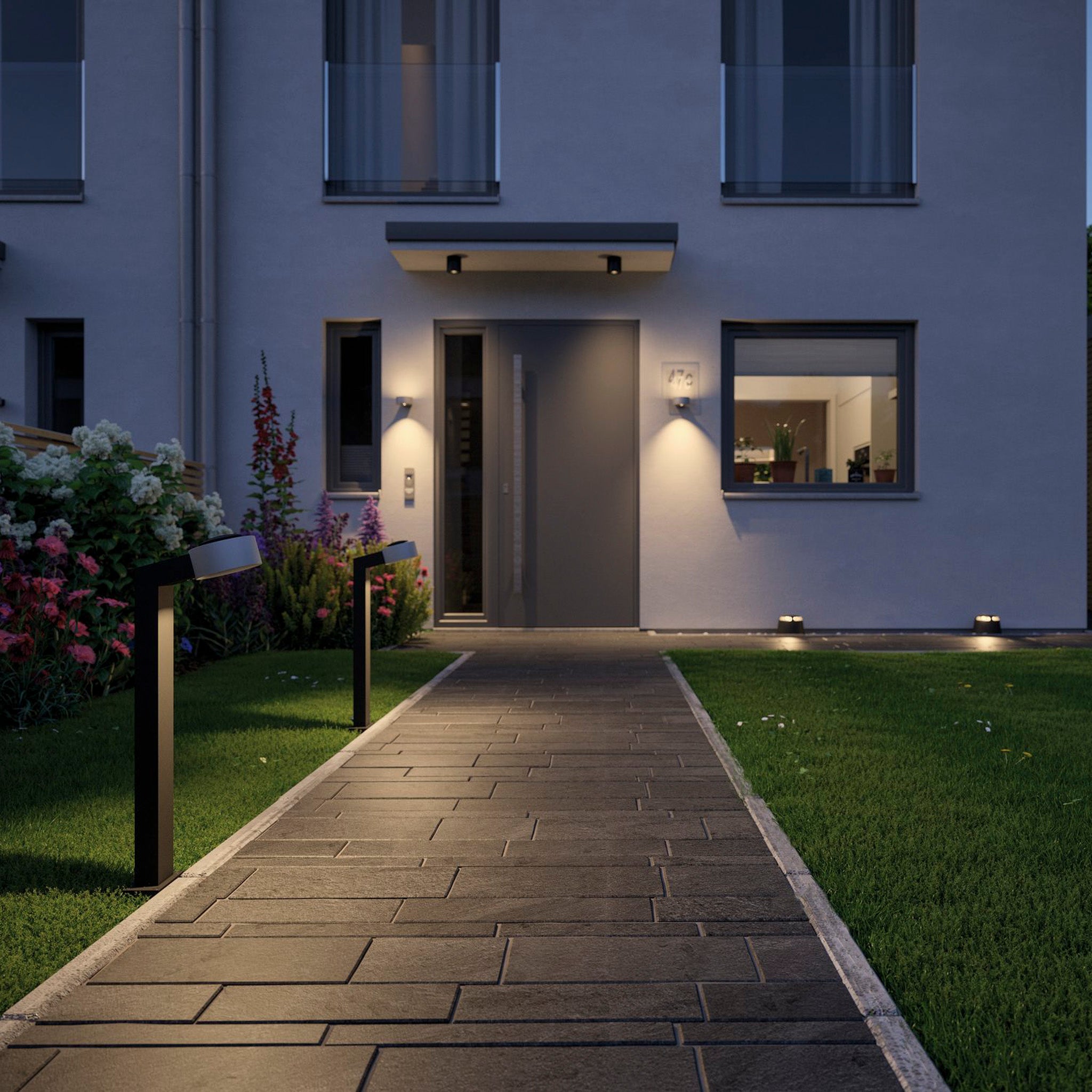 The entrance of the contemporary house features a stone path lit by weather-resistant outdoor fixtures. At night, the garden comes alive with lighting that highlights the lush grass and vibrant flowers. The dependable Paulmann - Plug & Shine Outdoor 6W LED Surface-Mounted 19.3cm Double Spotlight in Anthracite ensures a captivating ambiance every evening.