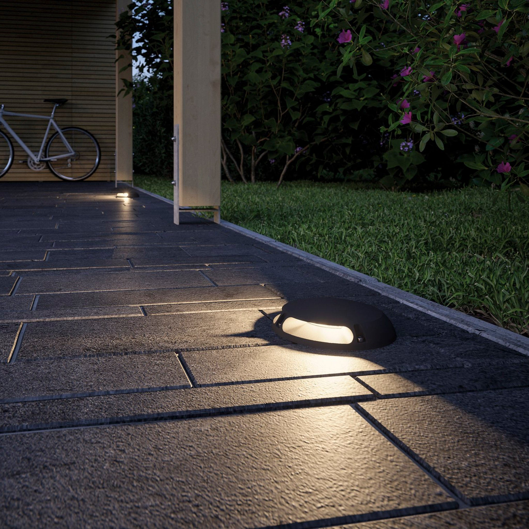 The tiled pathway beside the lush lawn is illuminated by the weather-resistant Paulmann Plug & Shine Outdoor 3.3W LED Surface-Mounted 16cm Double Spotlight in Anthracite, with a bicycle and vibrant flowers enhancing the background.