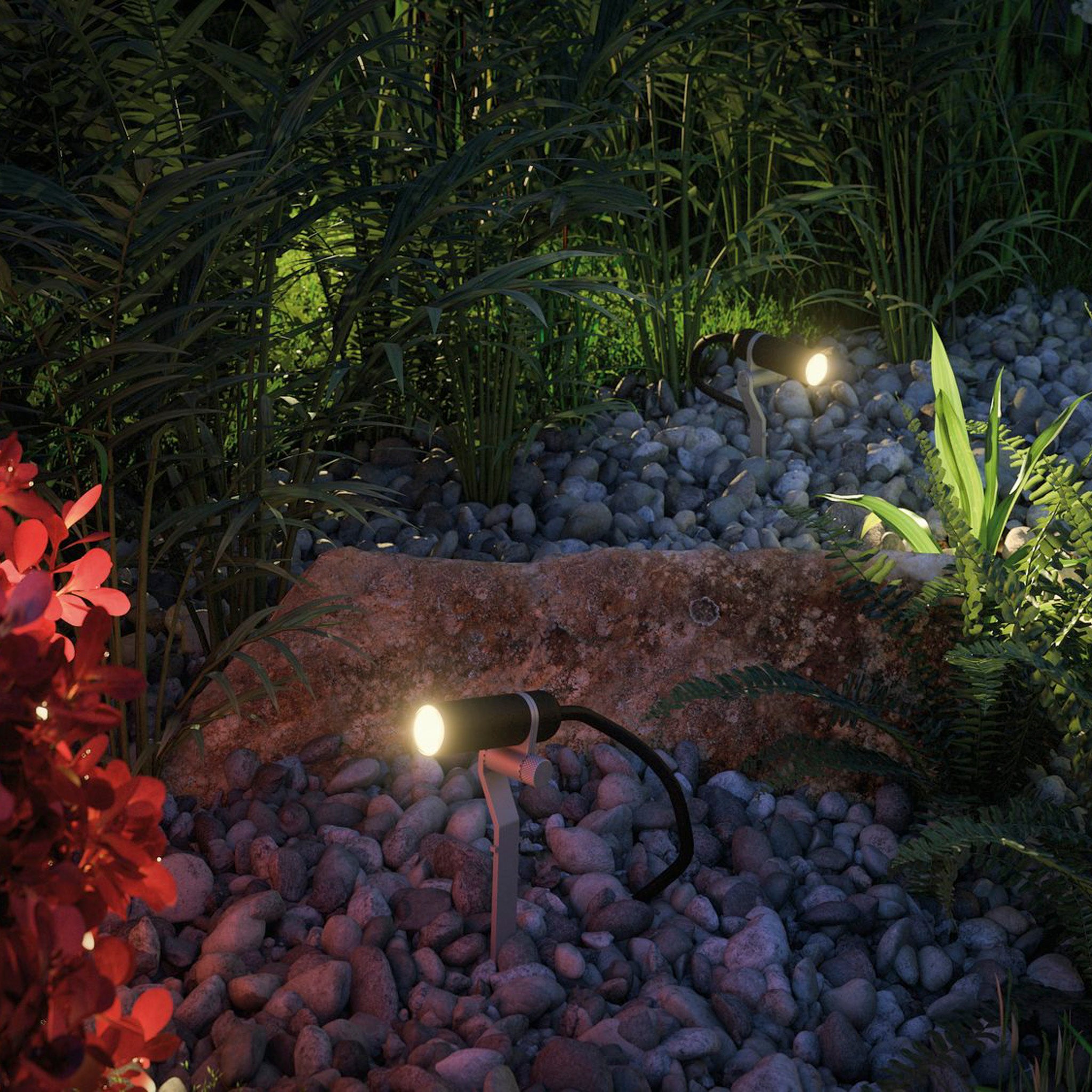 The Paulmann Plug & Shine Outdoor Plantini 2W LED Individual Spotlight in Anthracite illuminates plants and a rocky area at night, casting a warm white light that highlights the surrounding foliage and stones.