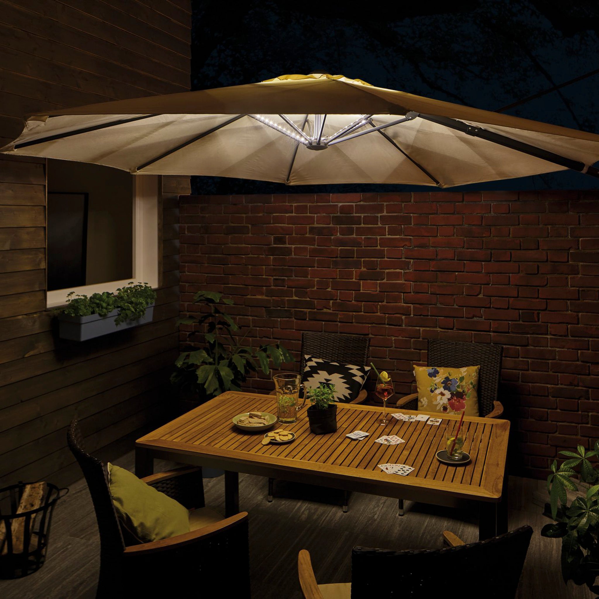 Under the starlit sky, the outdoor patio comes alive with a cozy gathering around a wooden table. Chairs are set invitingly under an illuminated umbrella adorned with the Paulmann - Outdoor LED 4x40cm Parasol Strips in Black, enhancing the warm and enchanting atmosphere perfect for a night of cards.